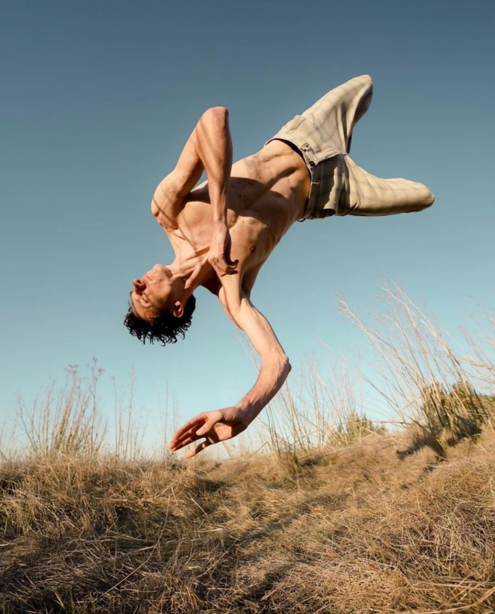 A person upside-down in a backbend, outdoors. Reaching arm, blue sky, dry grass.