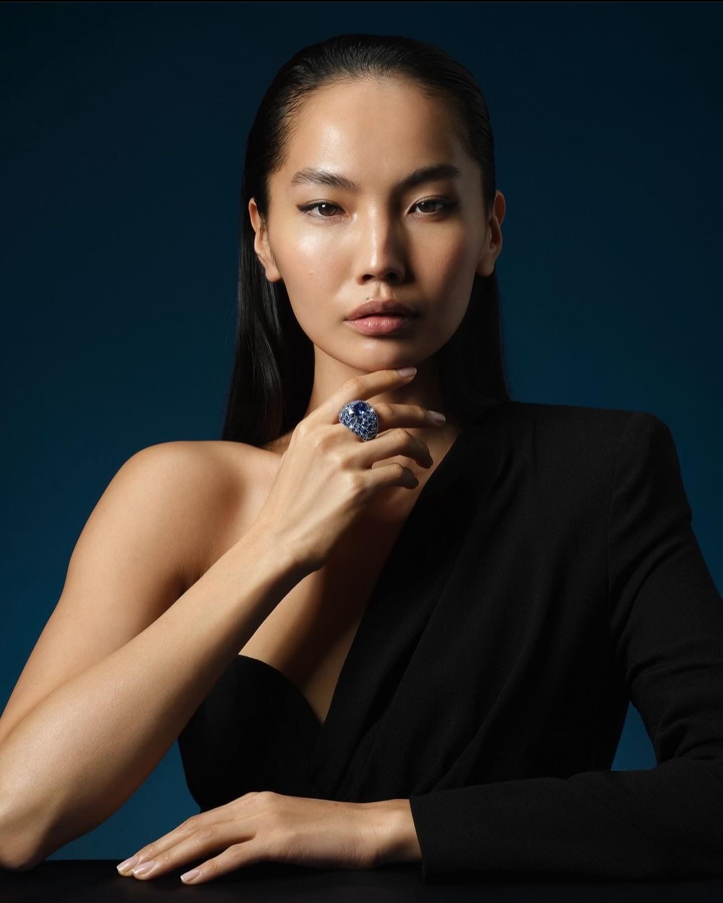 Woman in black blazer, hand to chin, wearing large blue ring, against a blue background.