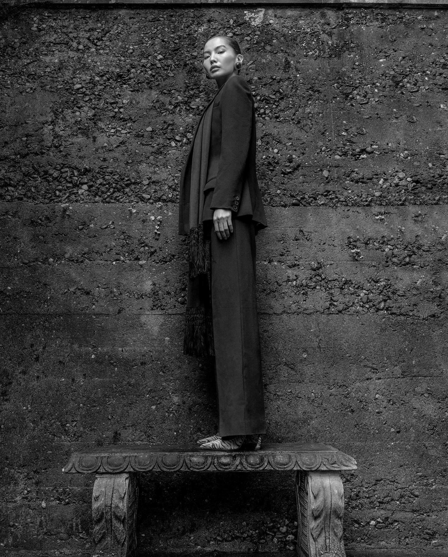 Woman in a suit stands on a stone bench in front of a textured wall.