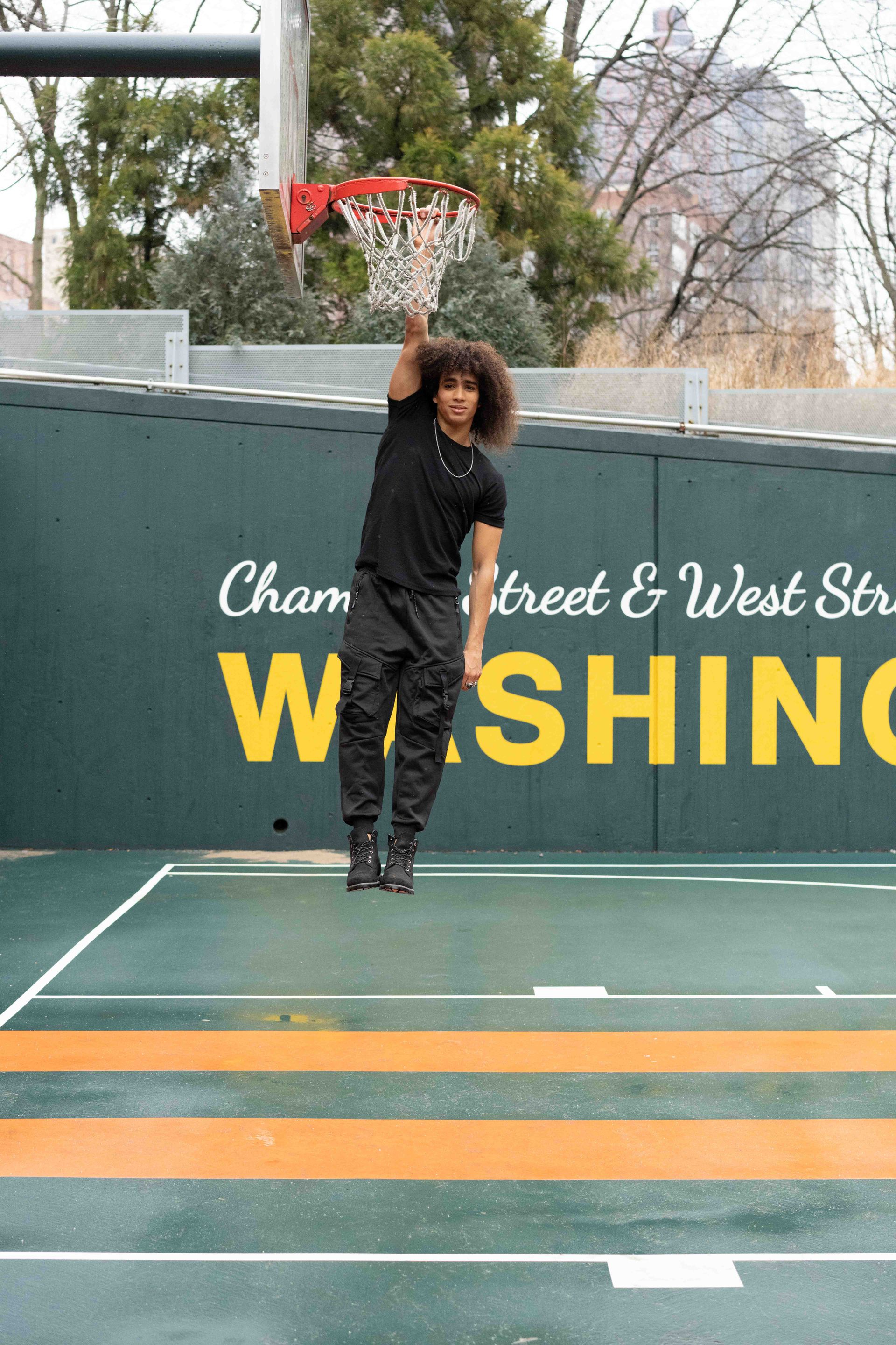 Man stretching on basketball court, leaning to the side, wearing black clothing. Court has green and orange lines.