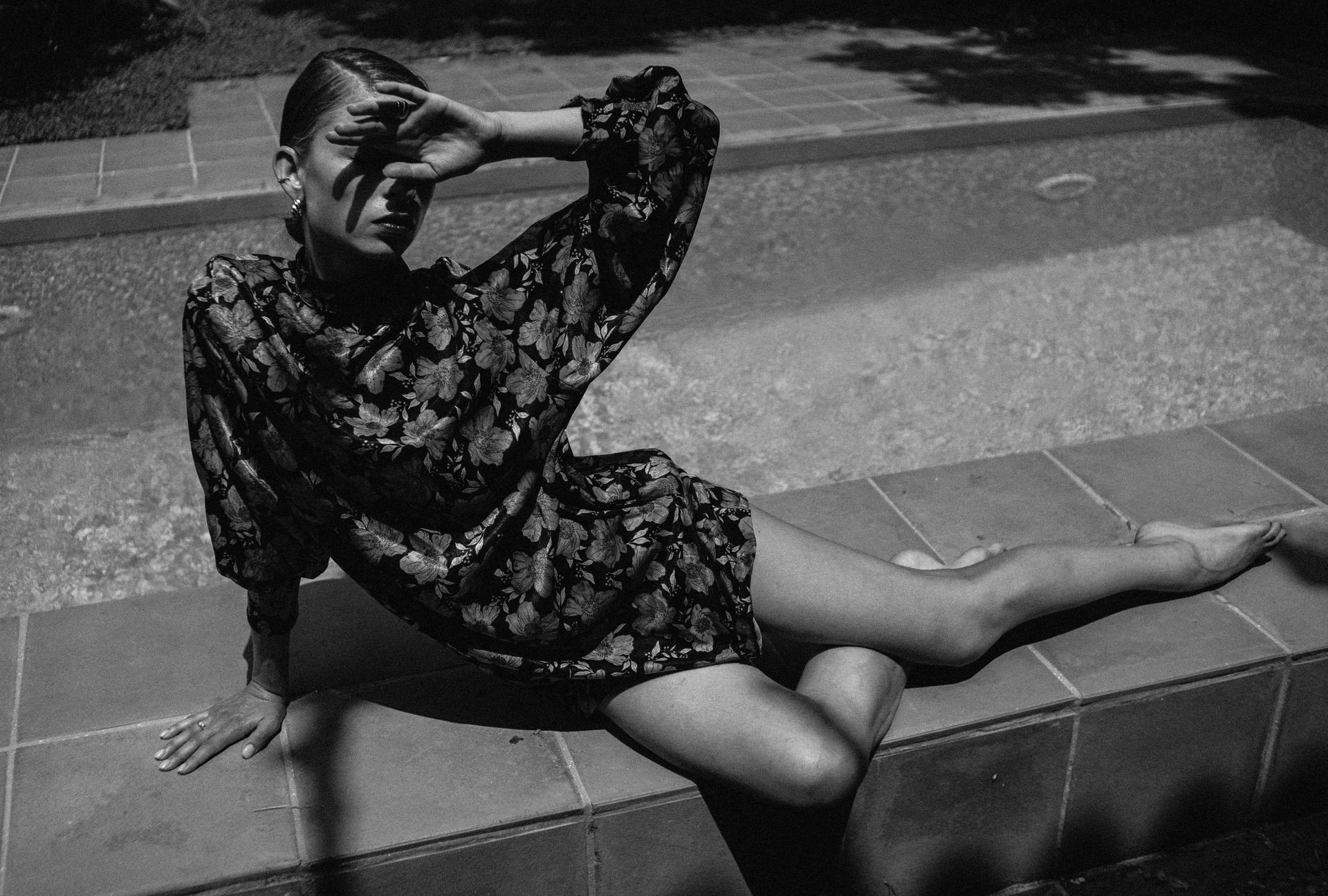 Woman in floral dress, shielding eyes from sun, lounging by a pool.