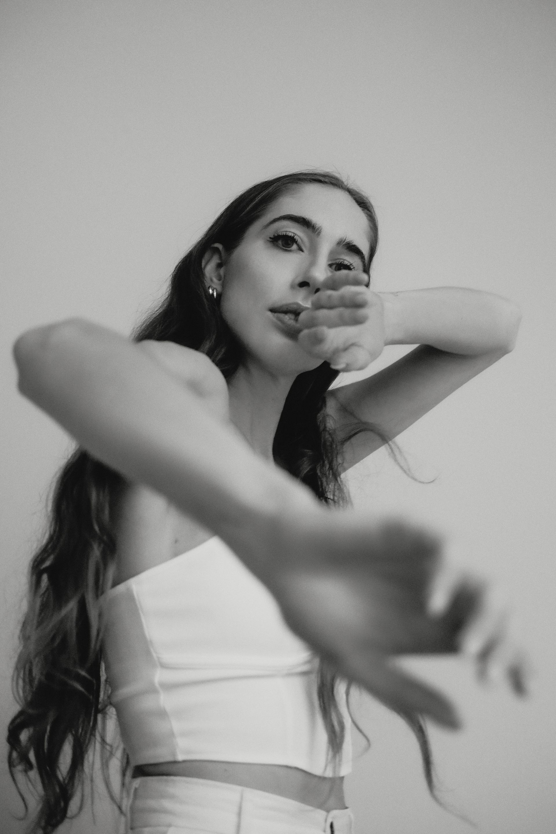 Woman in a white top, hand over mouth, reaching forward. Long hair, blurred background.