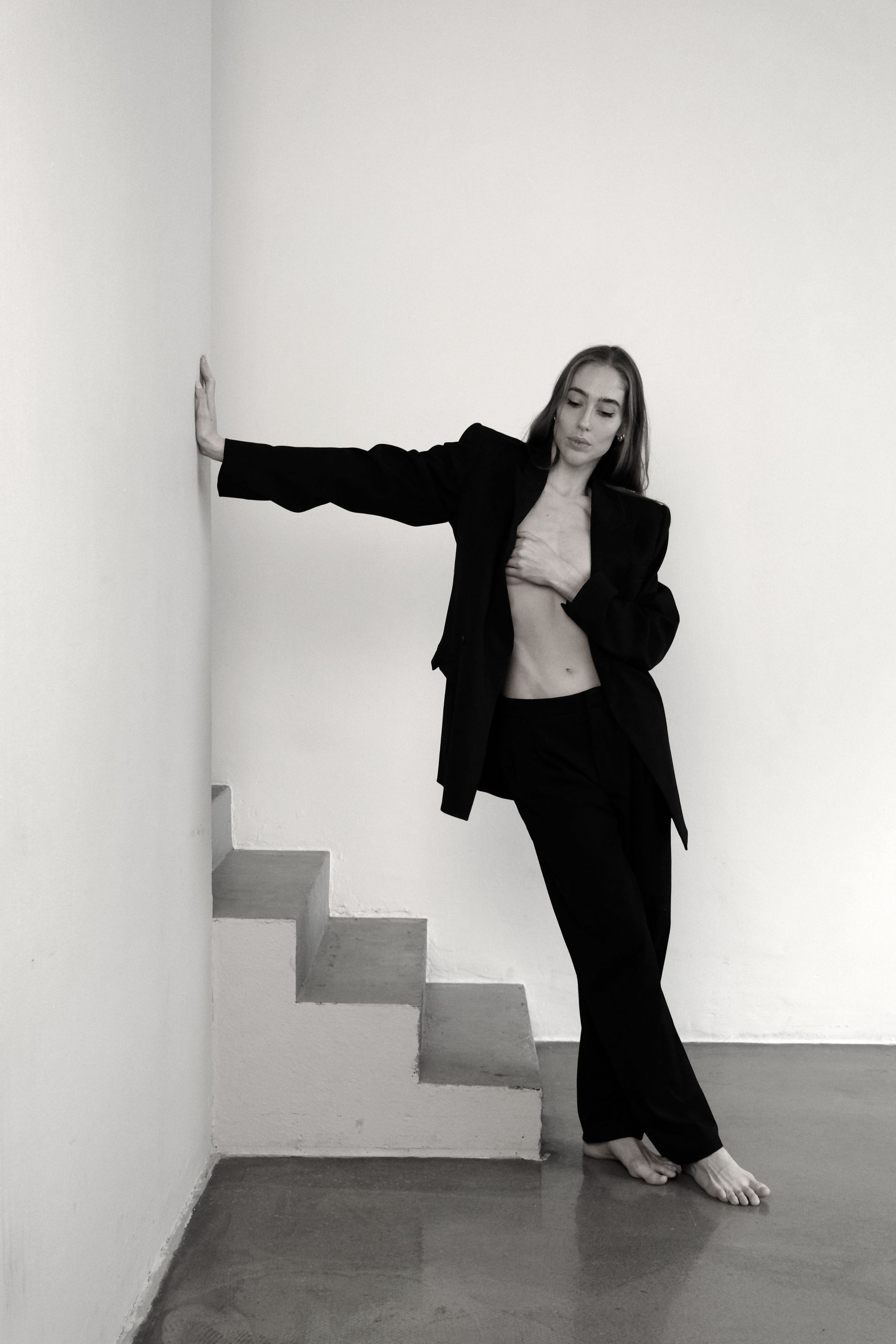 Woman in black suit leans against wall, partially baring chest. Staircase in background. Black and white.