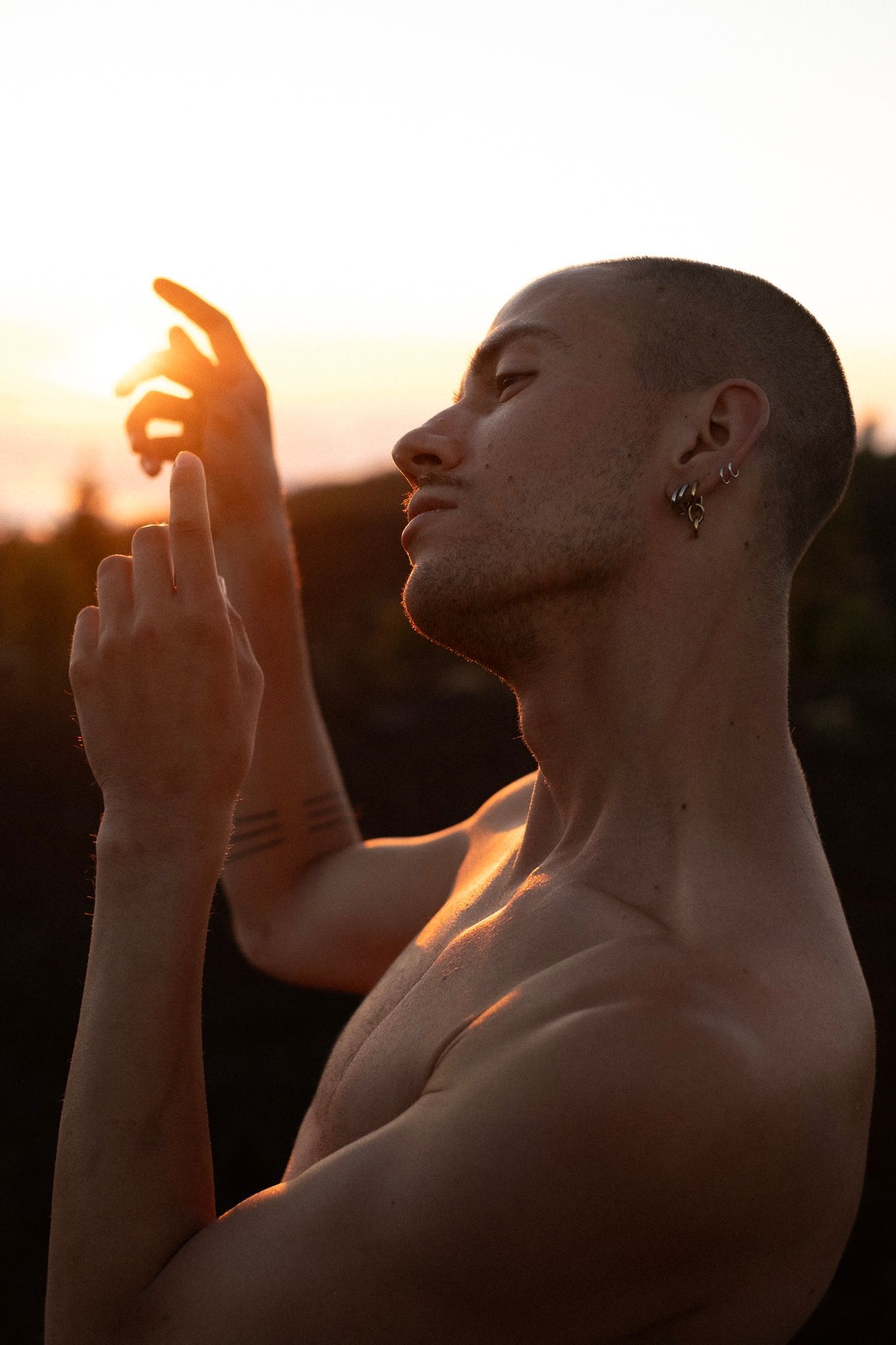 Man with shaved head reaching toward the sun; sunset illuminates his face and shoulders.