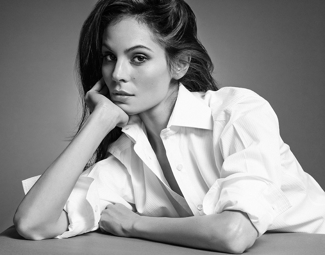 Woman in a white button-down shirt, resting chin on hand, looking at camera.