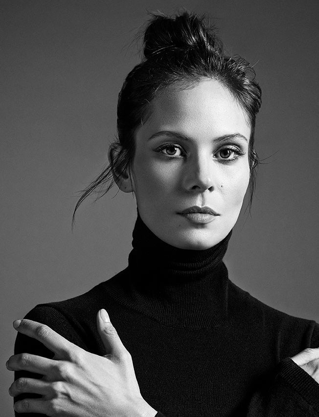 Woman with dark hair in a bun, wearing a black turtleneck, arms crossed, looking at the camera.