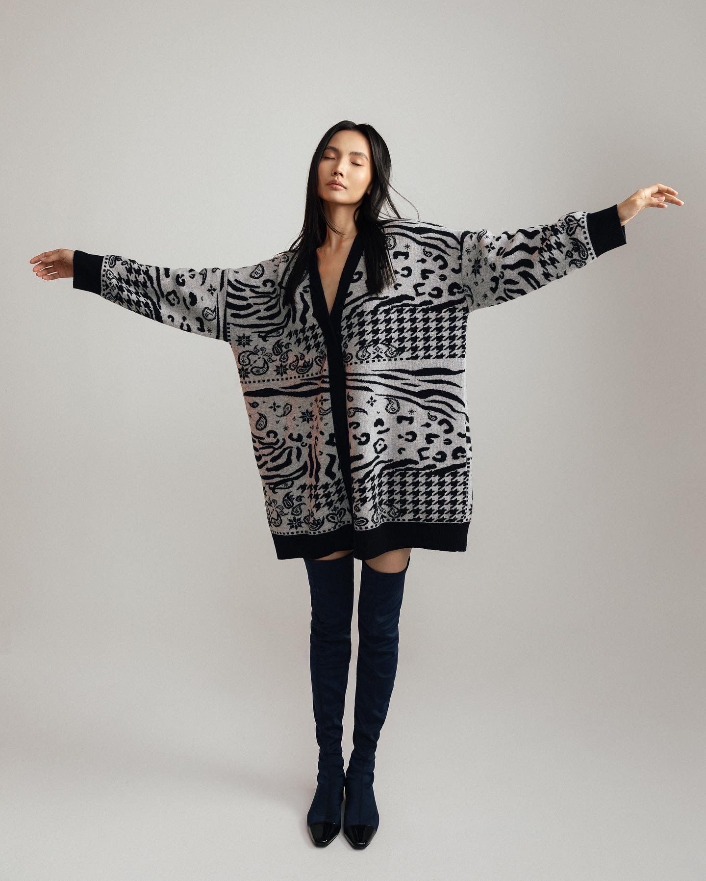Woman in patterned cardigan and thigh-high boots, arms outstretched.