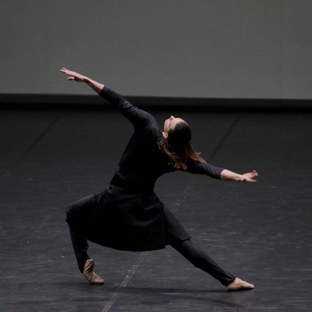 Dancer in black outfit performs a graceful pose on a stage. One leg bent, arms outstretched.