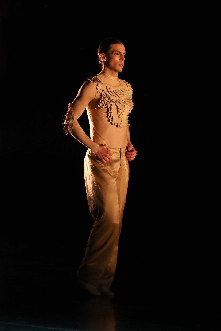 Man in beige, decorative top and pants, standing, hands on hips, in dark setting.