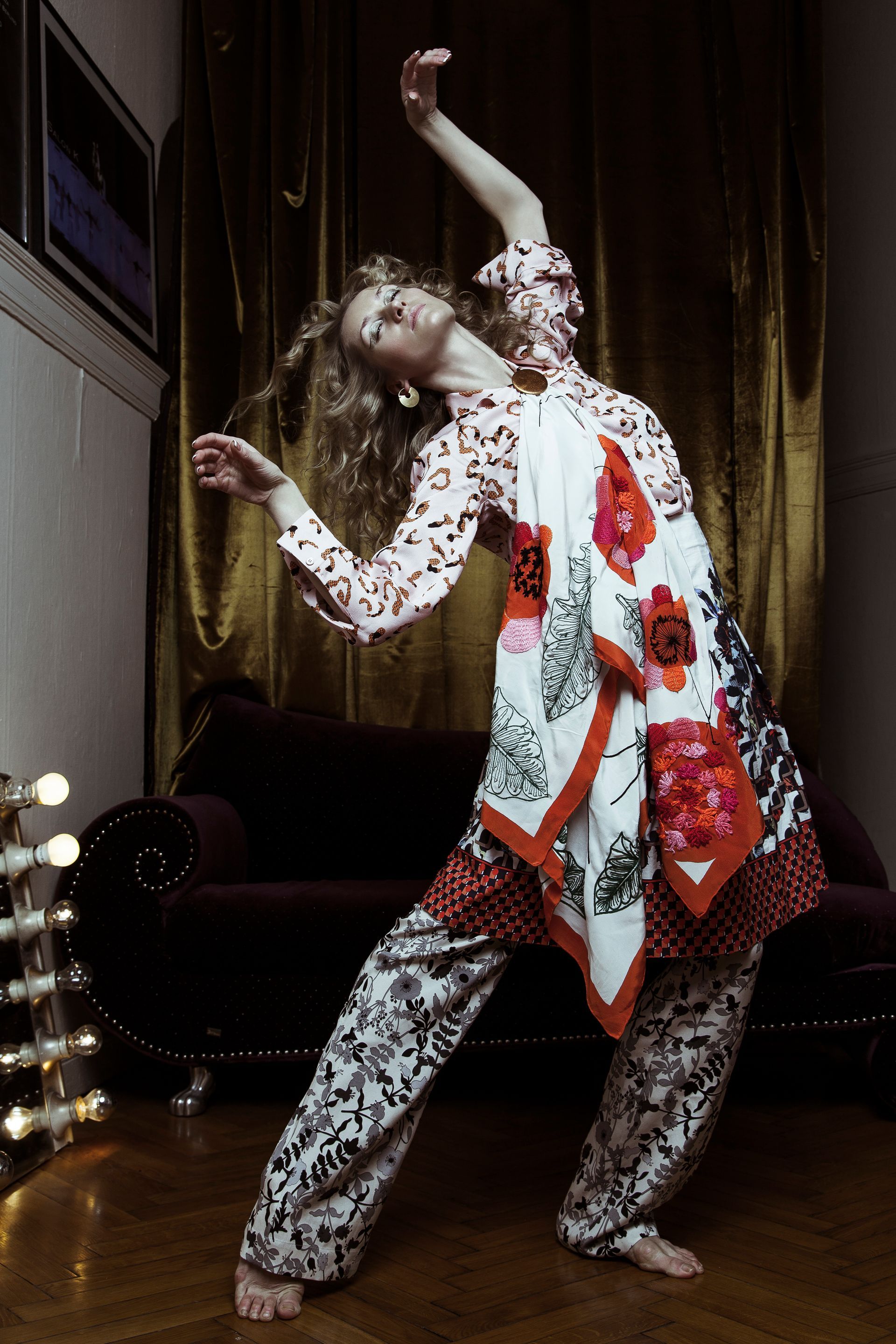 Woman in patterned outfit dances indoors with arched back, arm raised. Dramatic lighting, dark red sofa, gold curtains.