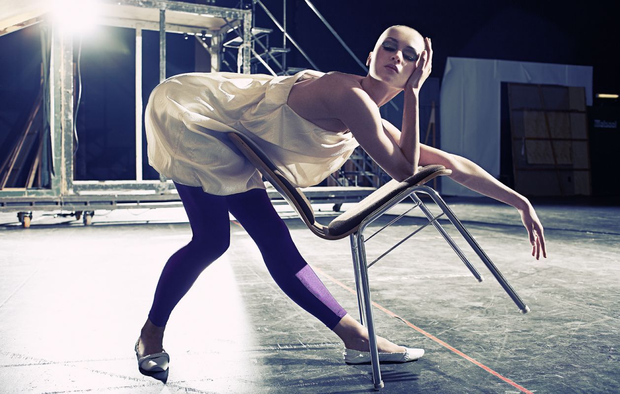 Woman in sequined dress and purple leggings leaning on chair, stage setting.