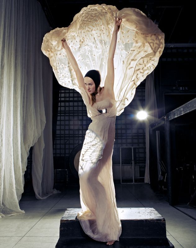 Woman in flowing white dress, arms raised, holding up a large, lacy fabric. Standing on a platform in a dark space.