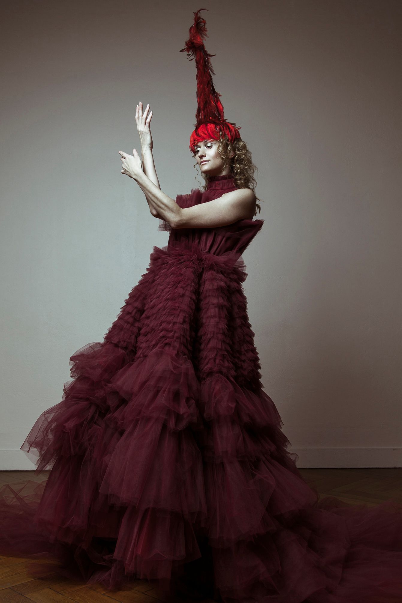 Woman in elaborate burgundy tulle gown and headdress, arms raised, in a room.