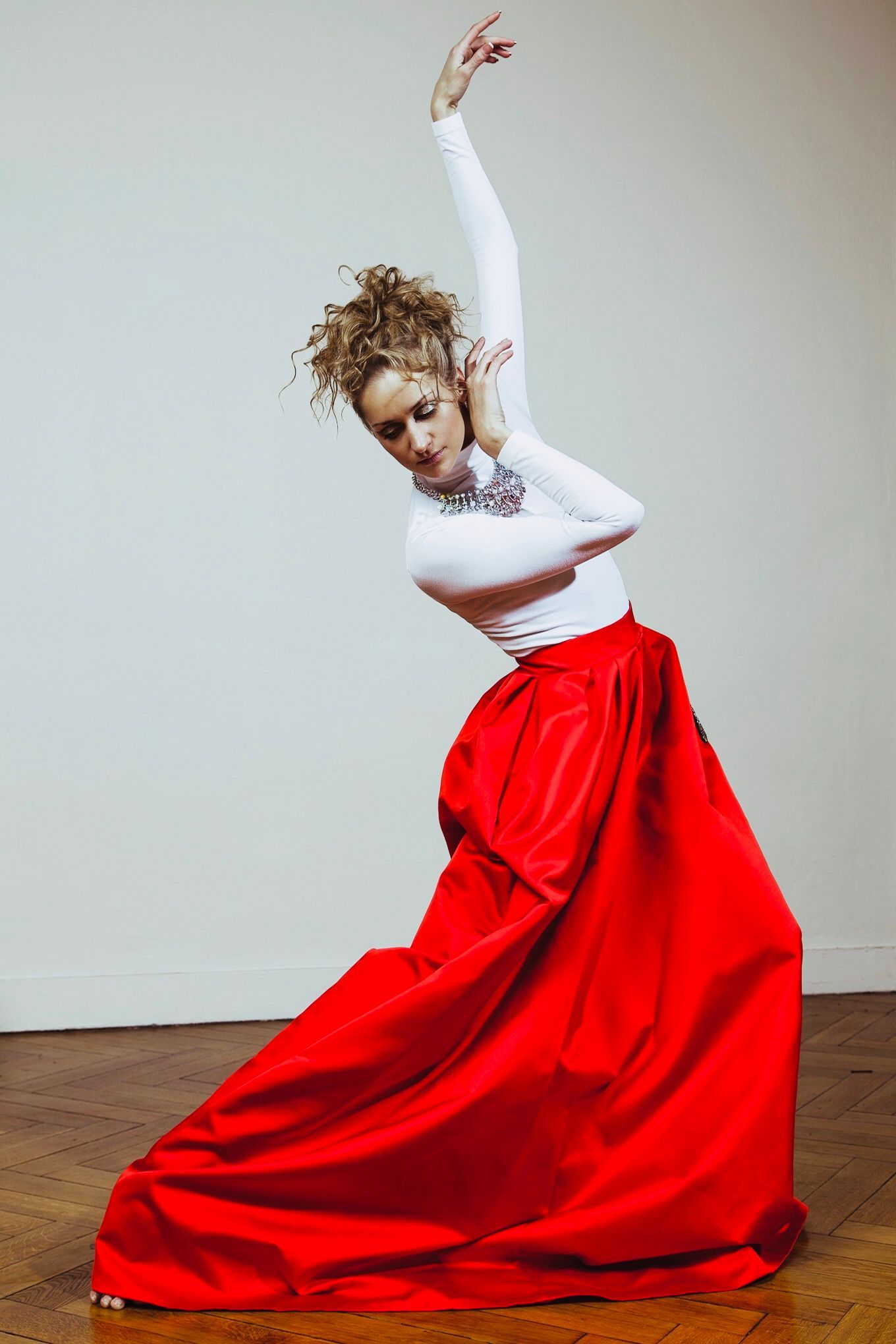 Woman in white top and red skirt poses in a dance. She arches, raising one arm. Wooden floor, neutral wall.