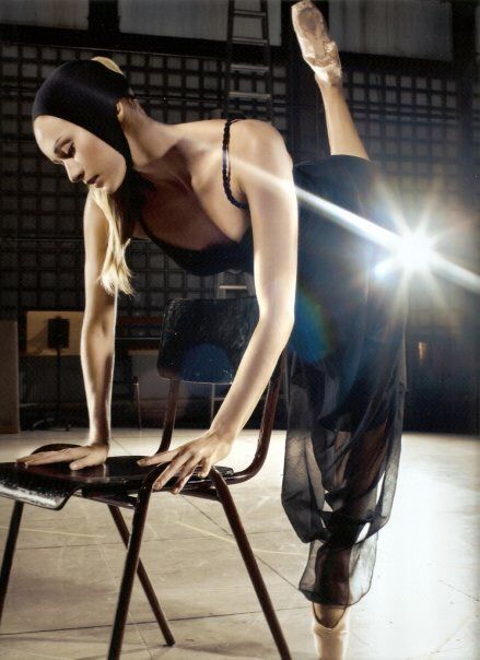 Ballerina in black outfit, using a chair for support, leg extended, indoors.