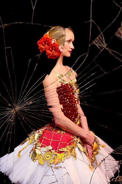 Ballerina in red and gold dress, hair with flowers, posed against shattered glass on black.