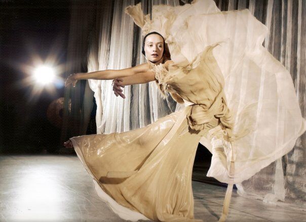 A woman in a flowing beige dress performs a ballet on stage. Bright light shines, arms outstretched.