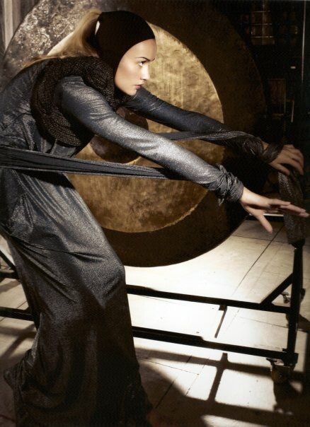 Woman in dark dress plays a large, golden gong in an indoor setting.