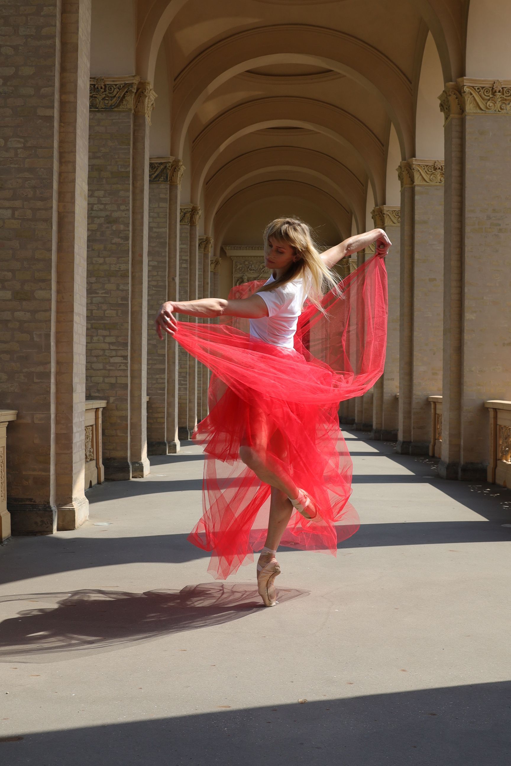 Ballerina in red tulle skirt and pointe shoes, dancing under an arched colonnade.