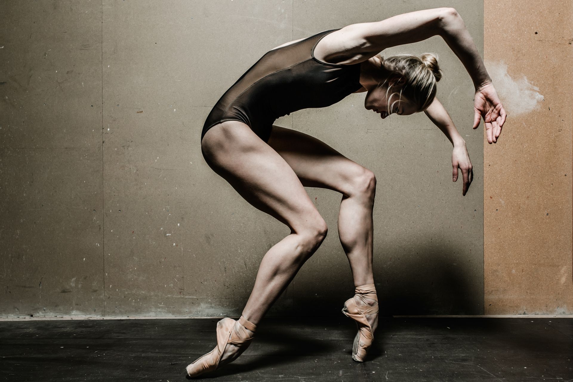 Ballerina in black leotard arches, balancing on pointe. Tan walls, dark floor.