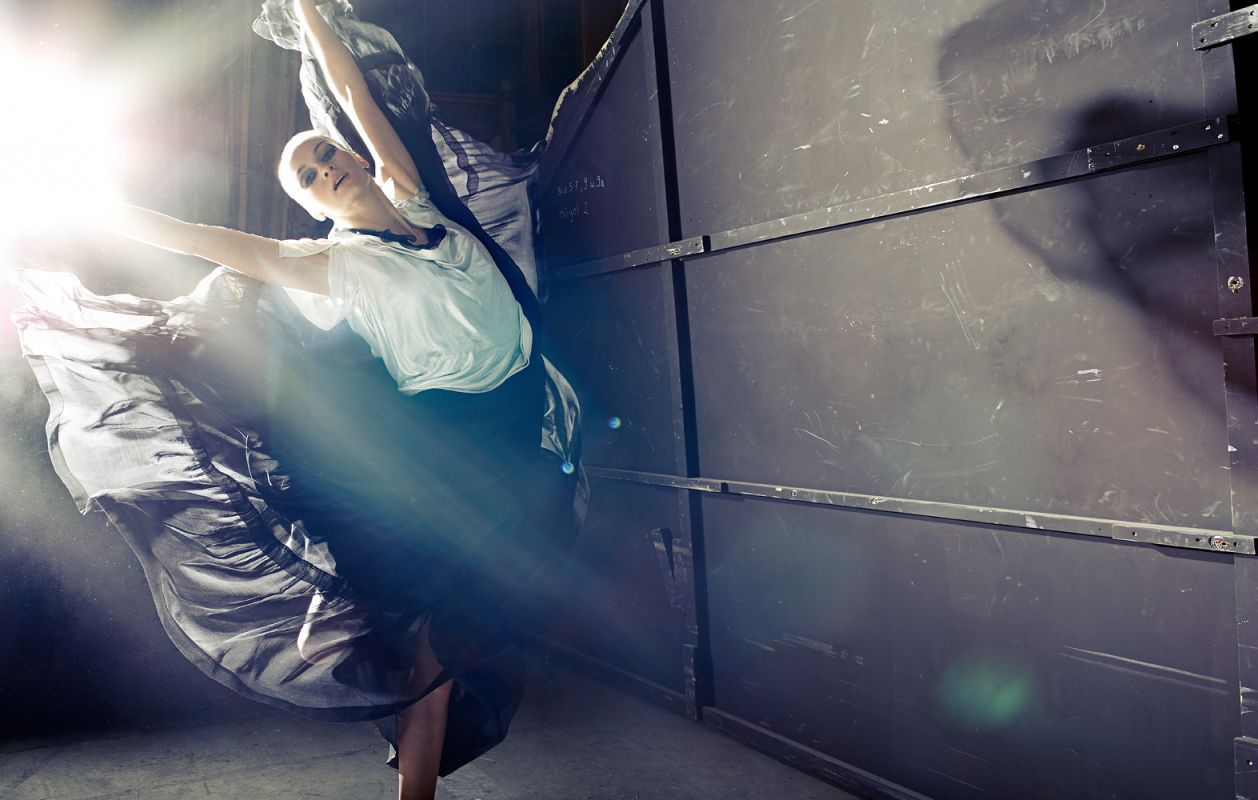 Ballet dancer in flowing skirt, arms outstretched, near a textured wall, with bright light shining.