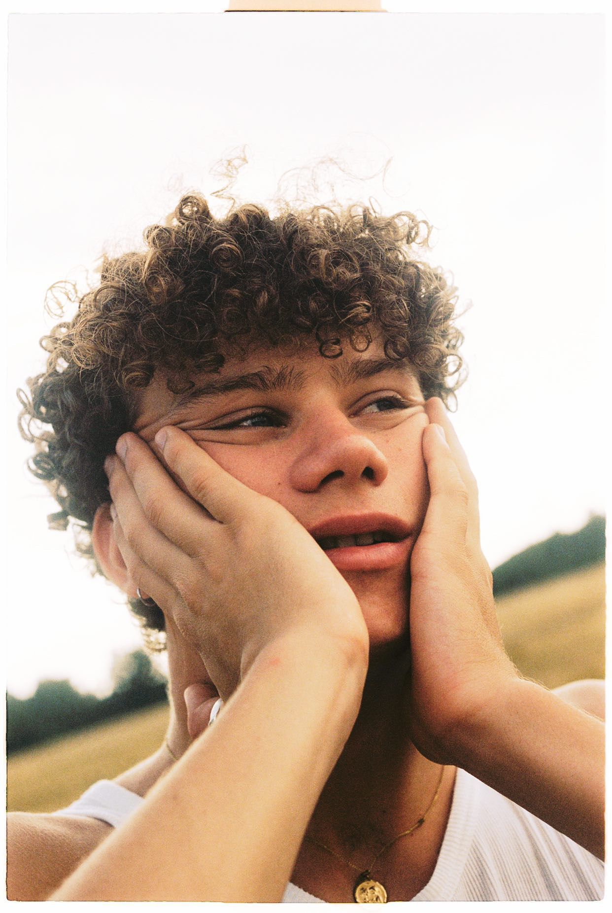 Person with curly hair, hands on cheeks, looking upward with a thoughtful expression; outdoors, neutral background.