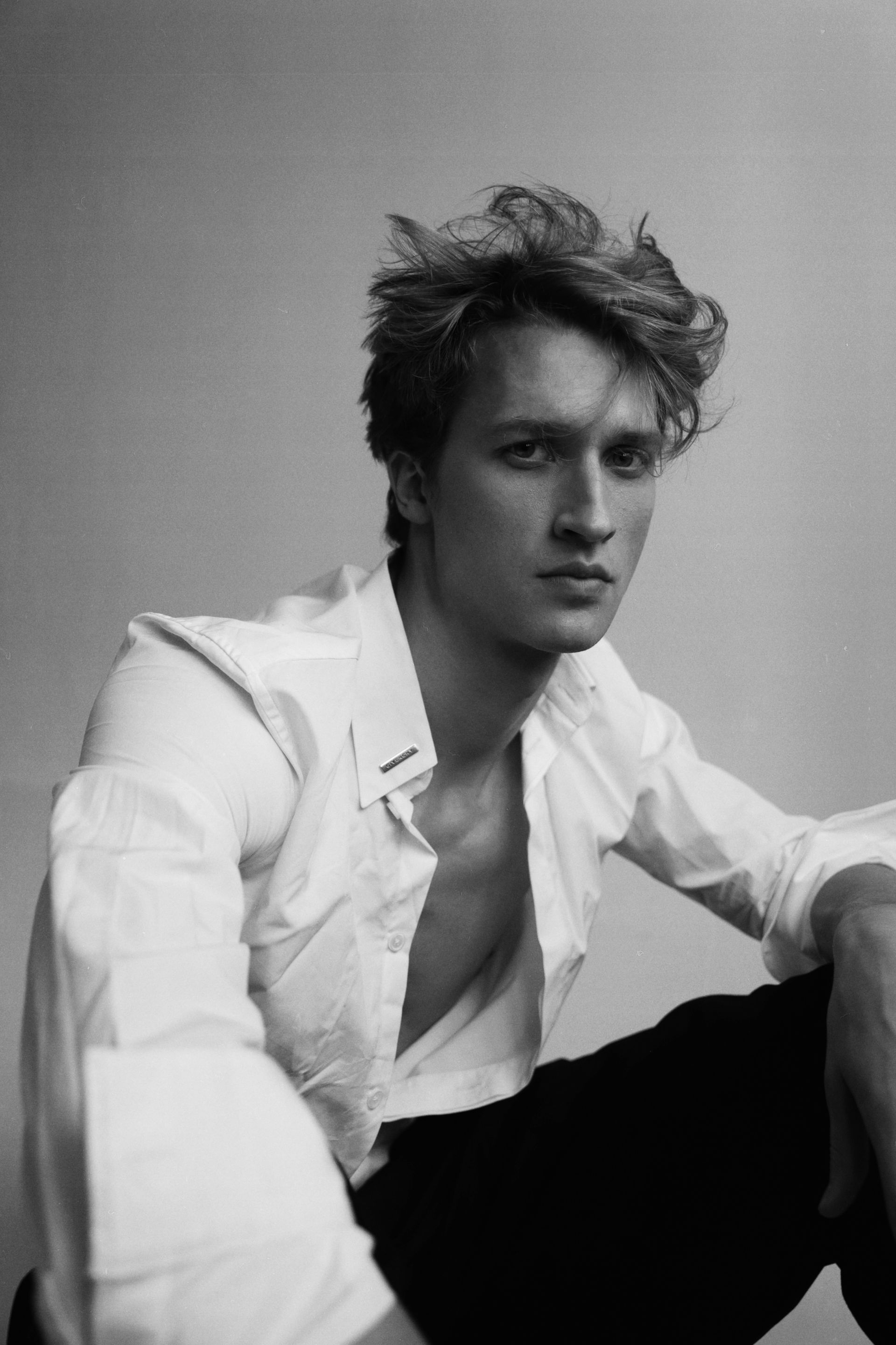 Man in white shirt, unbuttoned, looking at camera. Leaning forward, black pants. Black and white.