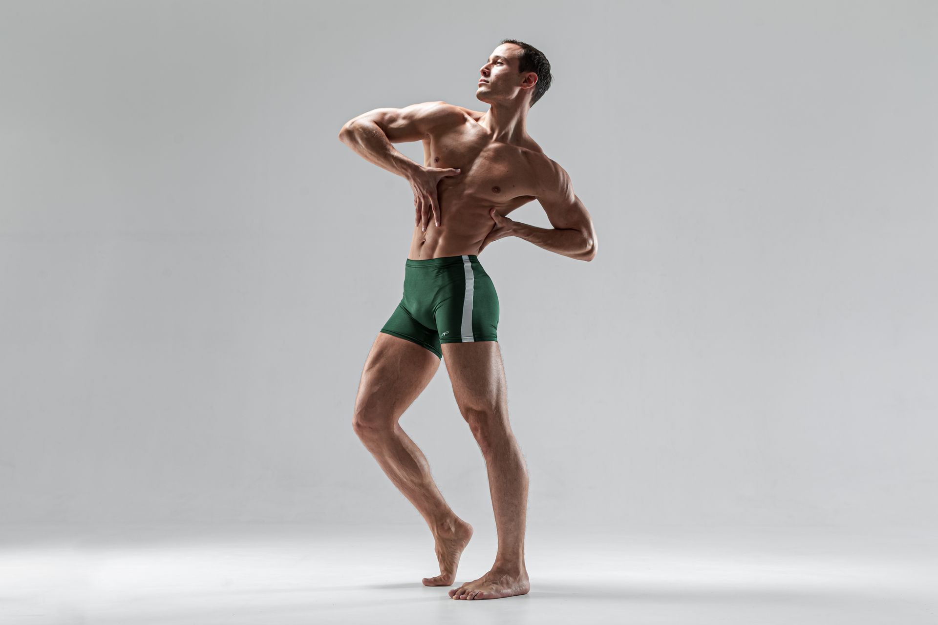 Fit person poses shirtless in green shorts, arching back, hands on chest. Bare feet, neutral backdrop.