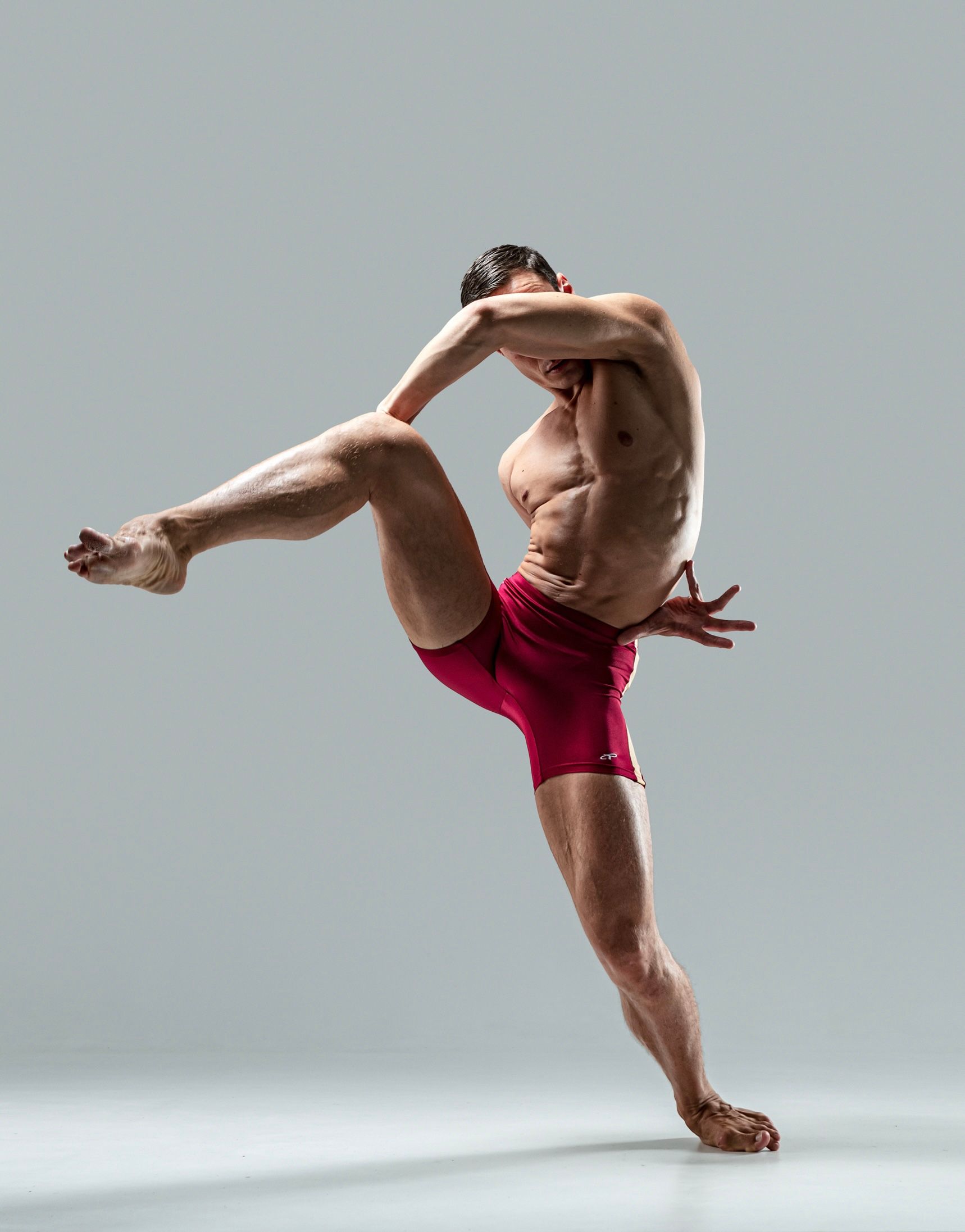 Male dancer in red shorts, mid-air pose, arms and leg bent, gray background.