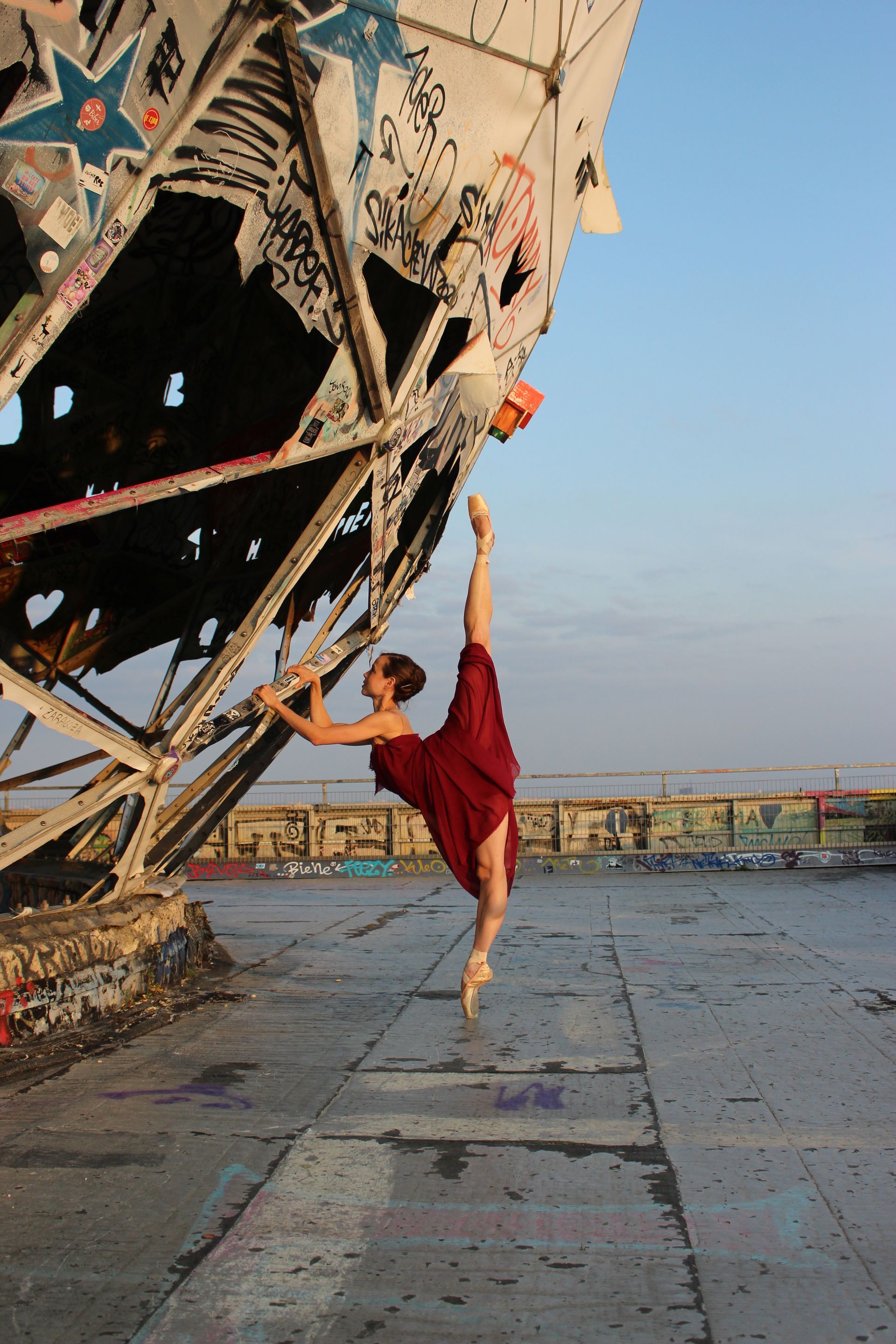Dancer in red dress performs a split, holding onto a graffiti-covered metal structure on a rooftop.