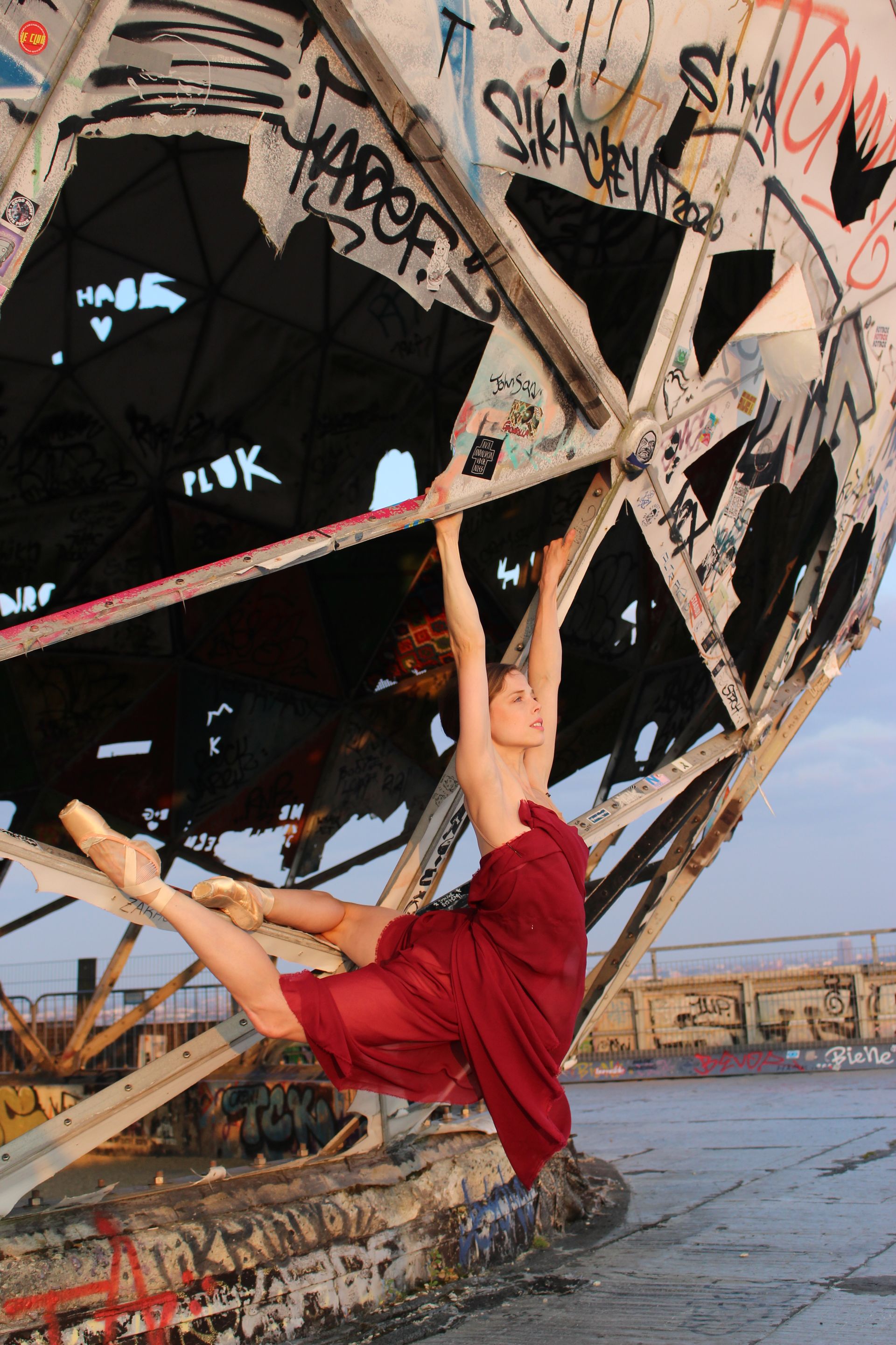 Ballerina in red dress hangs from graffiti-covered metal structure, legs extended, pointe shoes visible.