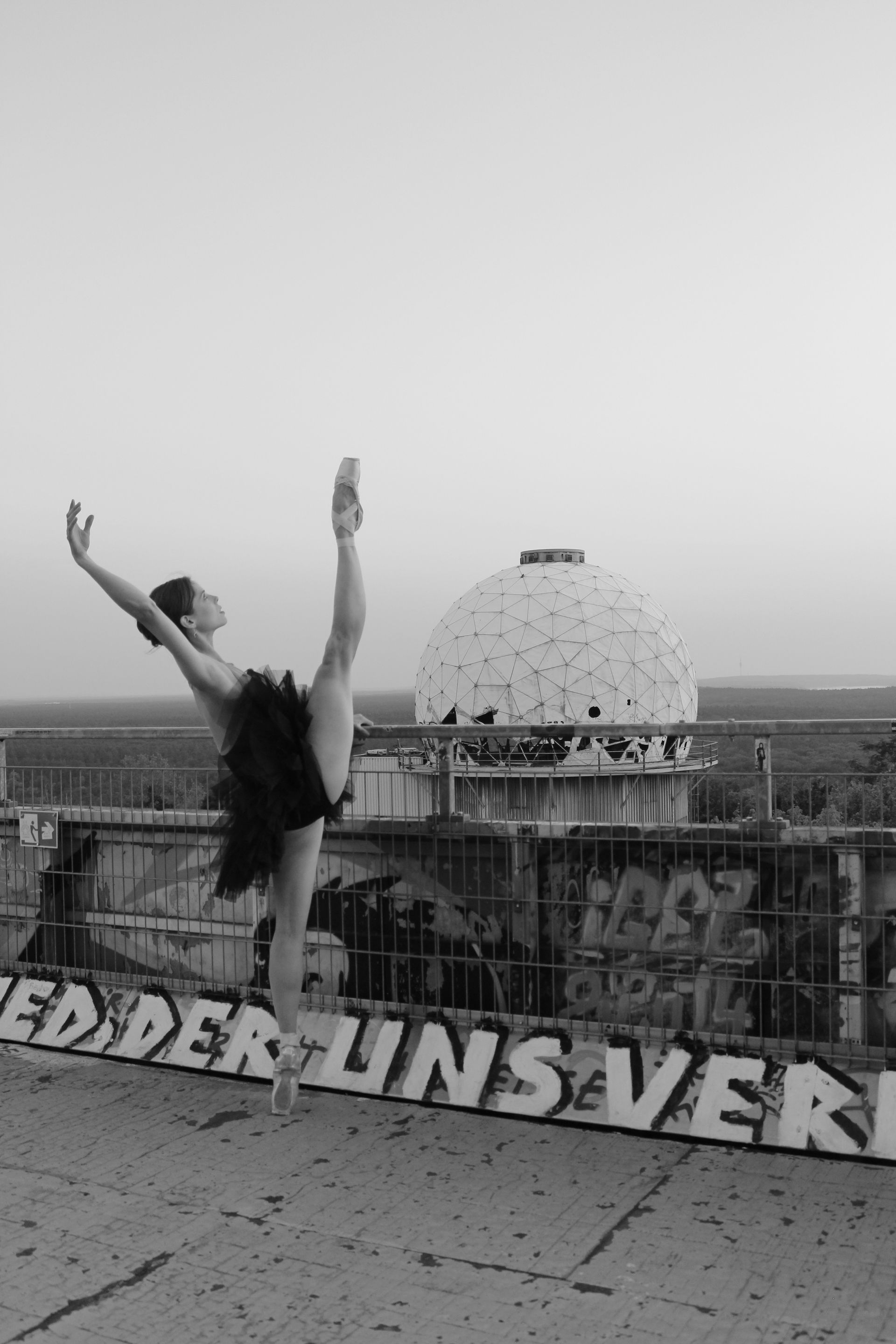 Ballet dancer in a black tutu balances on one leg, extending other leg upwards, on a rooftop with graffiti and dome.