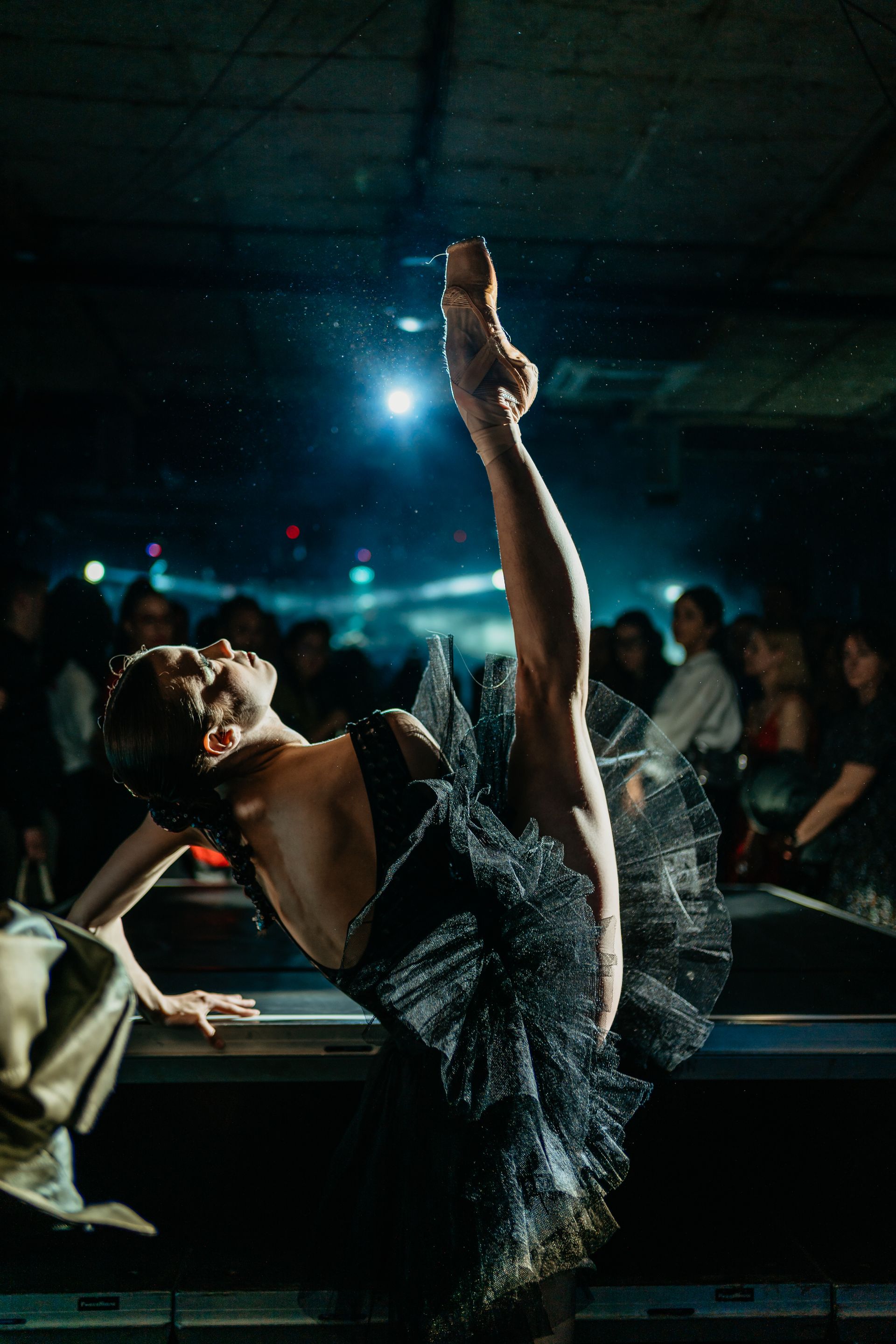 Ballet dancer in black tutu, leg extended high, performing on a stage with audience.