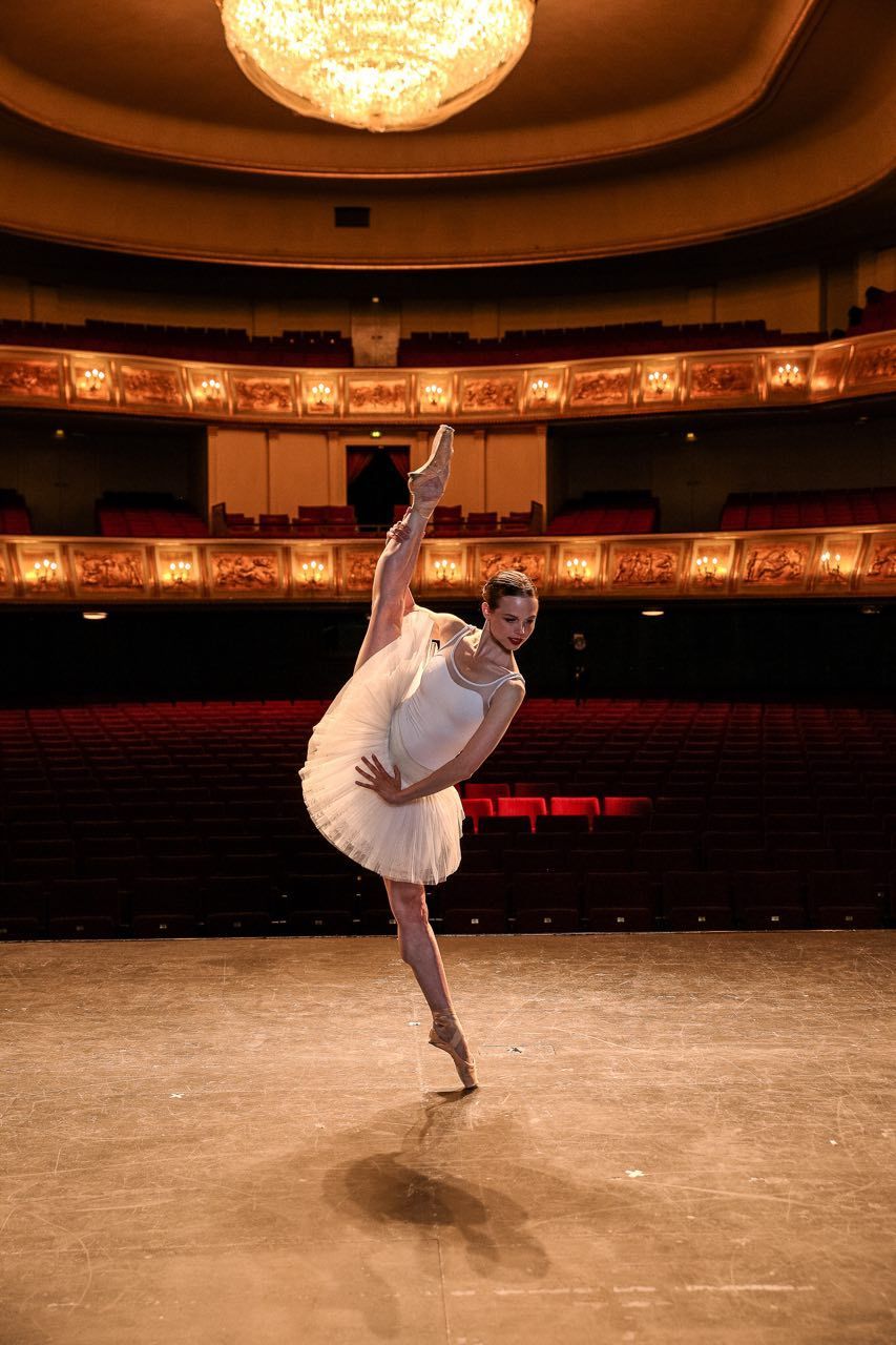 Ballerina in white tutu on stage, leg extended in a high split, illuminated by a chandelier. Red seating in background.