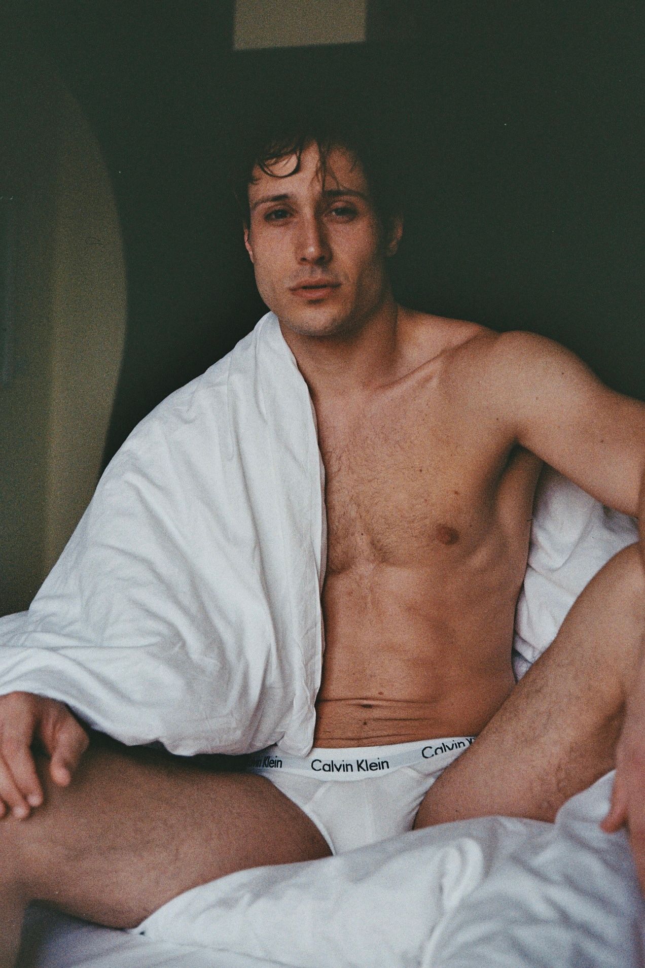 Man sitting, partially draped in a white towel, wearing white underwear, natural light.