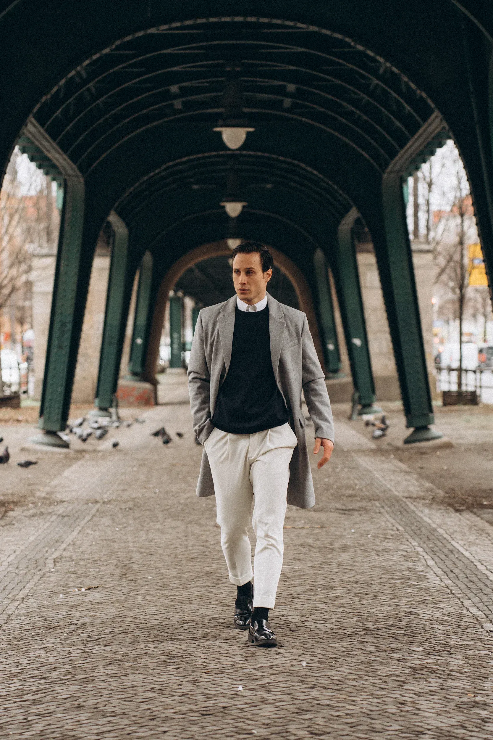 Man walks toward the camera under a covered walkway, wearing a long coat, sweater, and light pants.