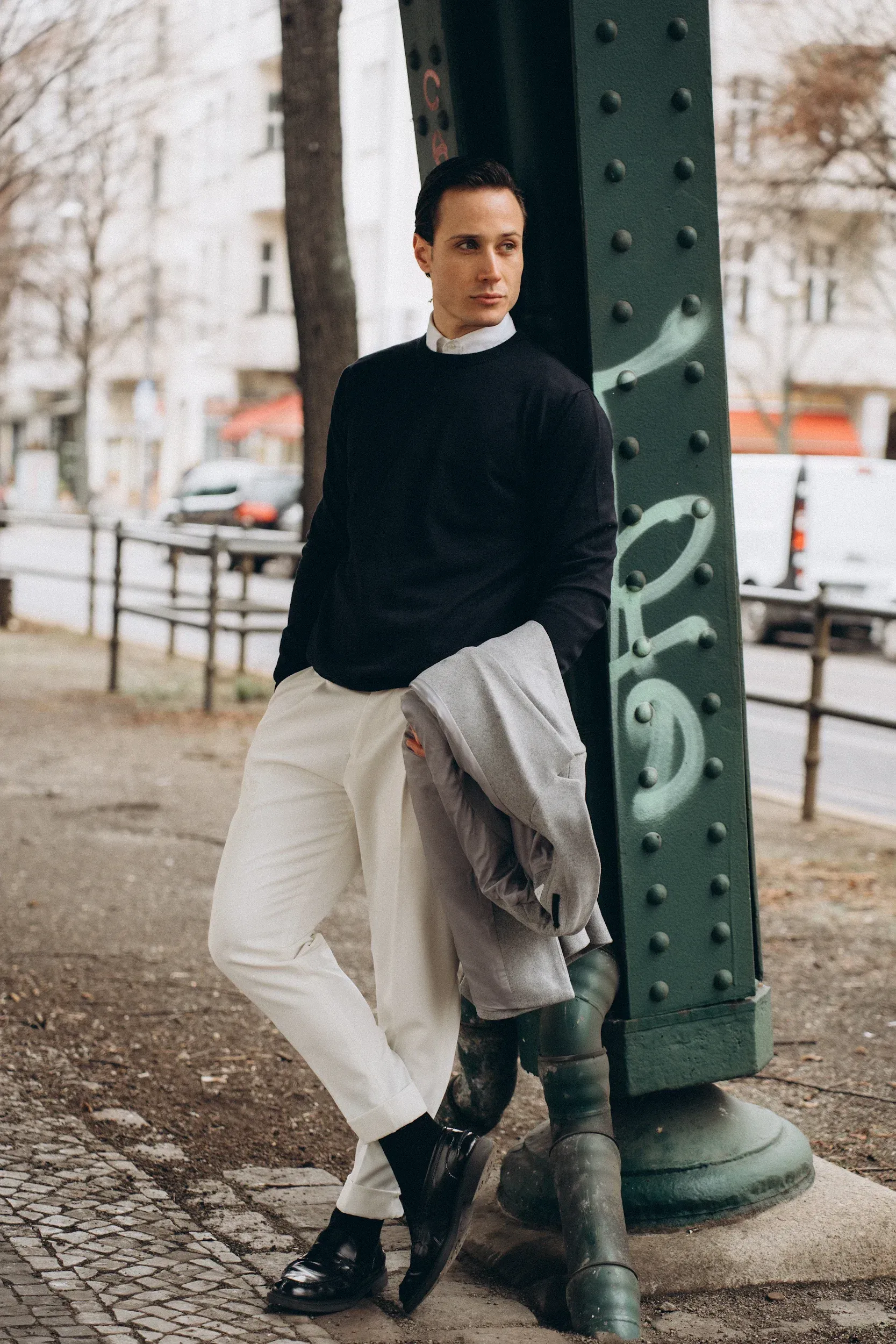 Man in black sweater, white pants leans on a green metal pole outdoors. Holds a gray jacket, looks away.