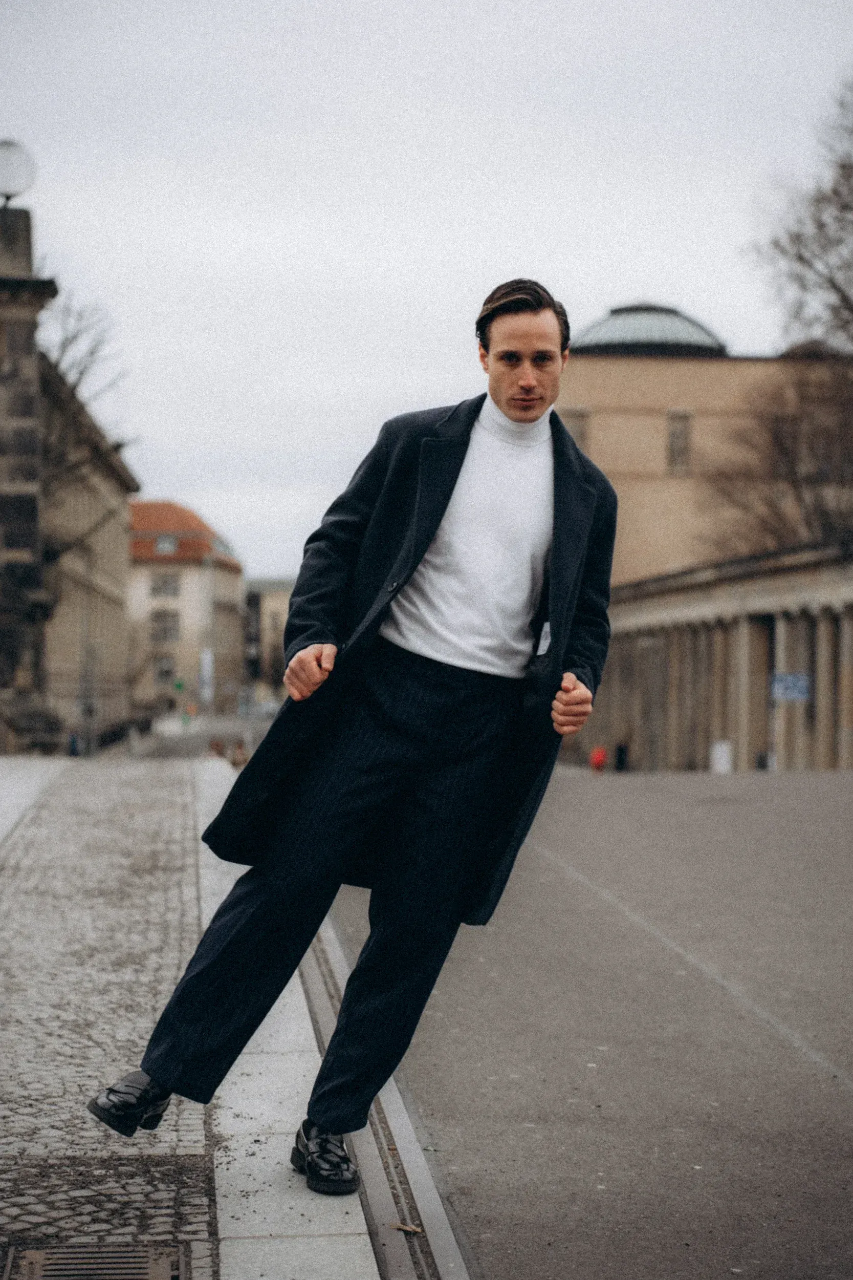 Man in dark coat and pants stands on curb in a city street, white turtleneck visible.