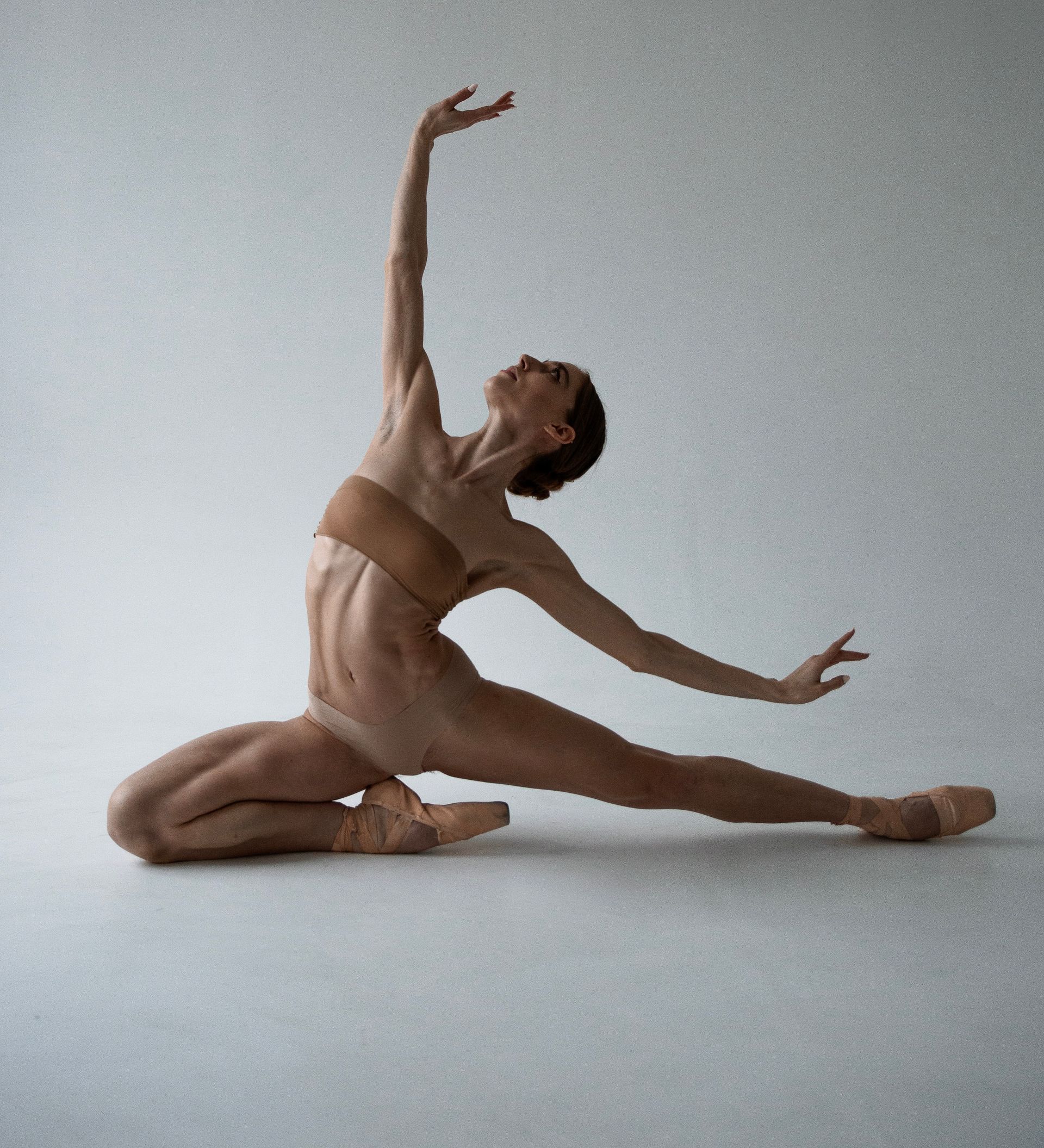 Ballet dancer in a split, arms extended. Leaning back on one knee, pale studio background.