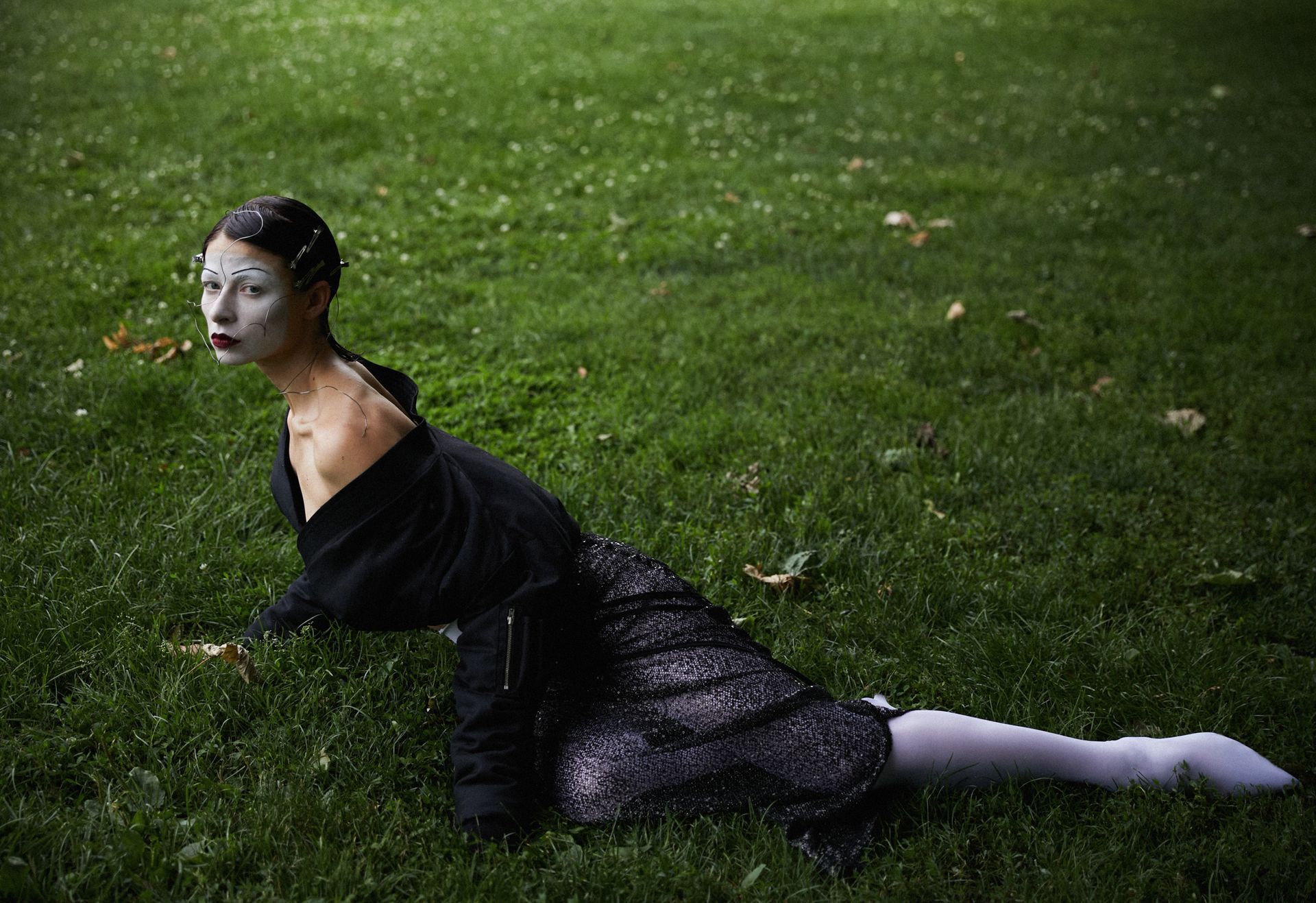 Woman in black clothing and white face makeup kneels on green grass.