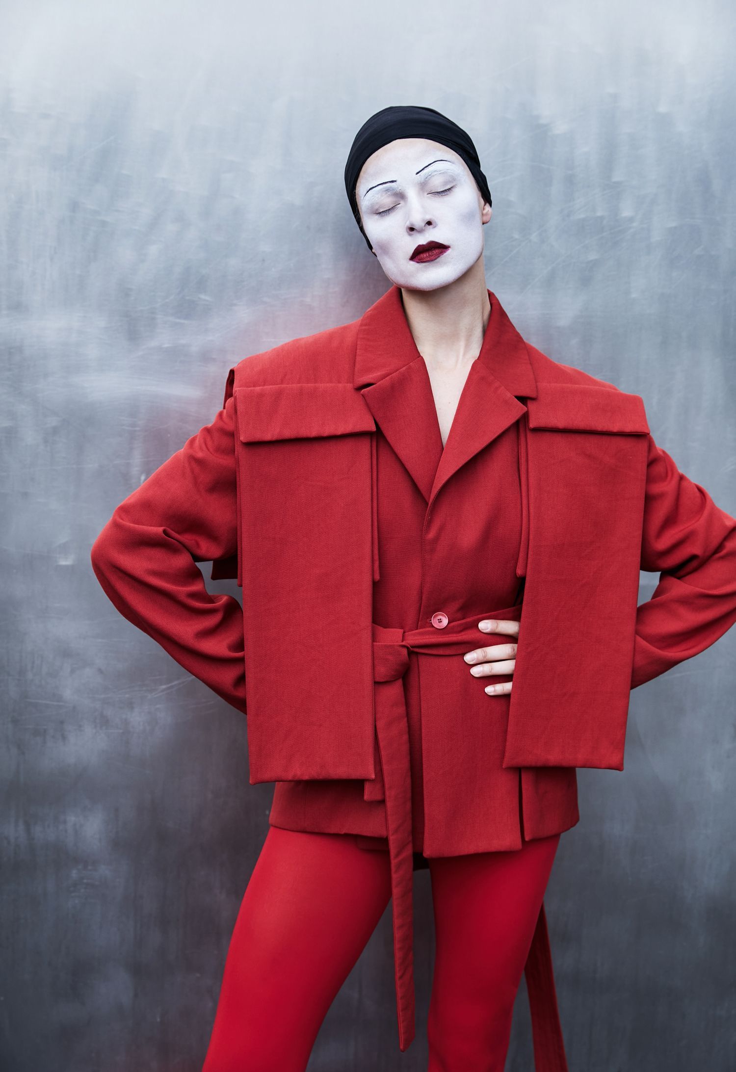 Person with white face paint and red lips in a red suit, standing with hands on hips against a silver wall.