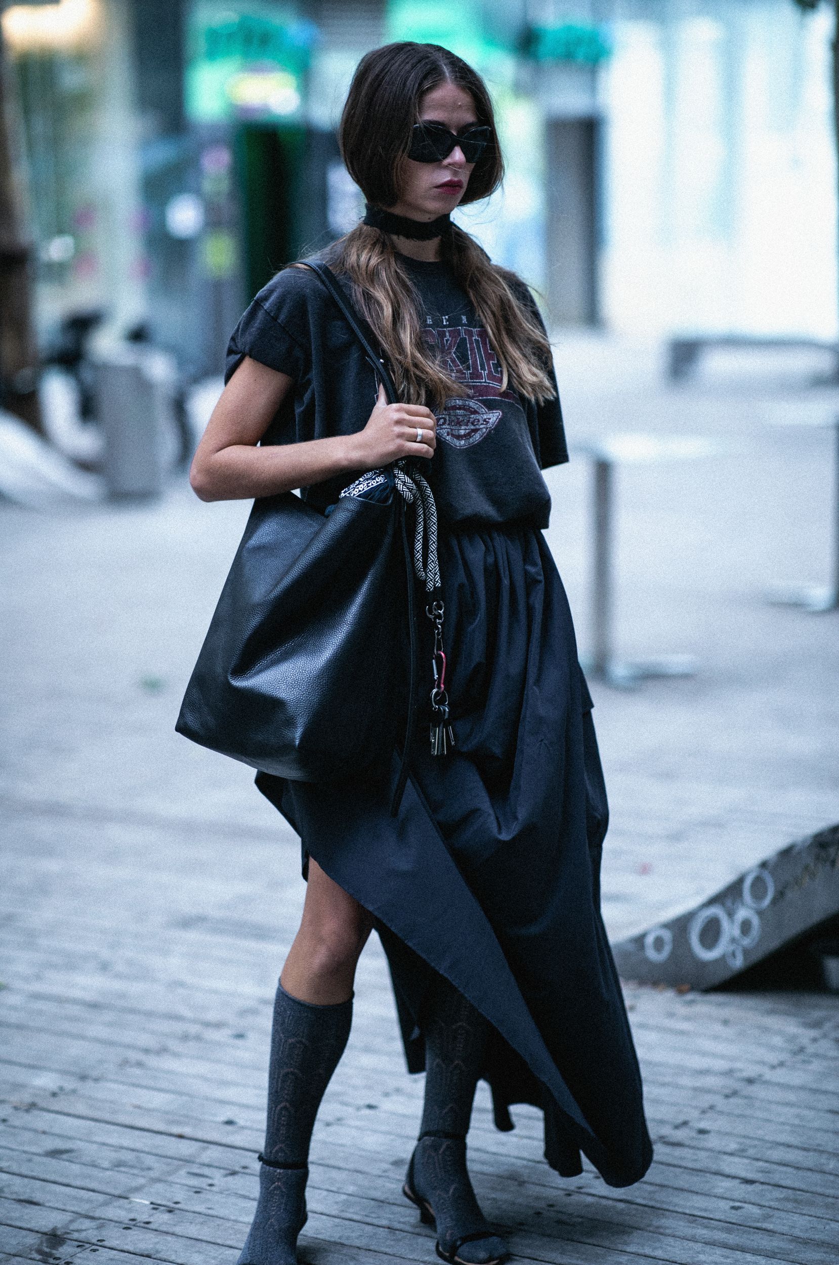 Woman in black outfit, sunglasses, carrying large bag, standing on street.