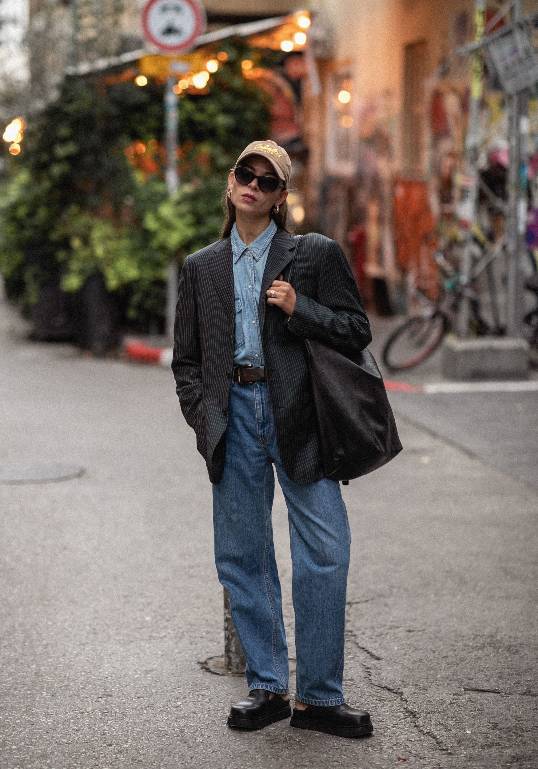 Person in oversized blazer, jeans, and cap stands on a city street.
