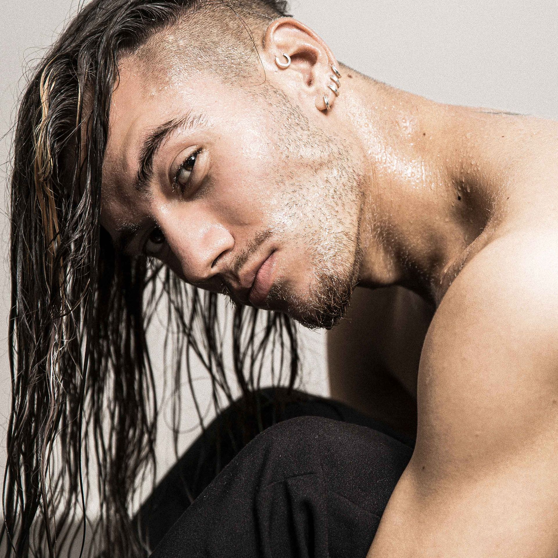 Man with wet hair, shaved sides, looking down; shirtless, dark pants, neutral background.
