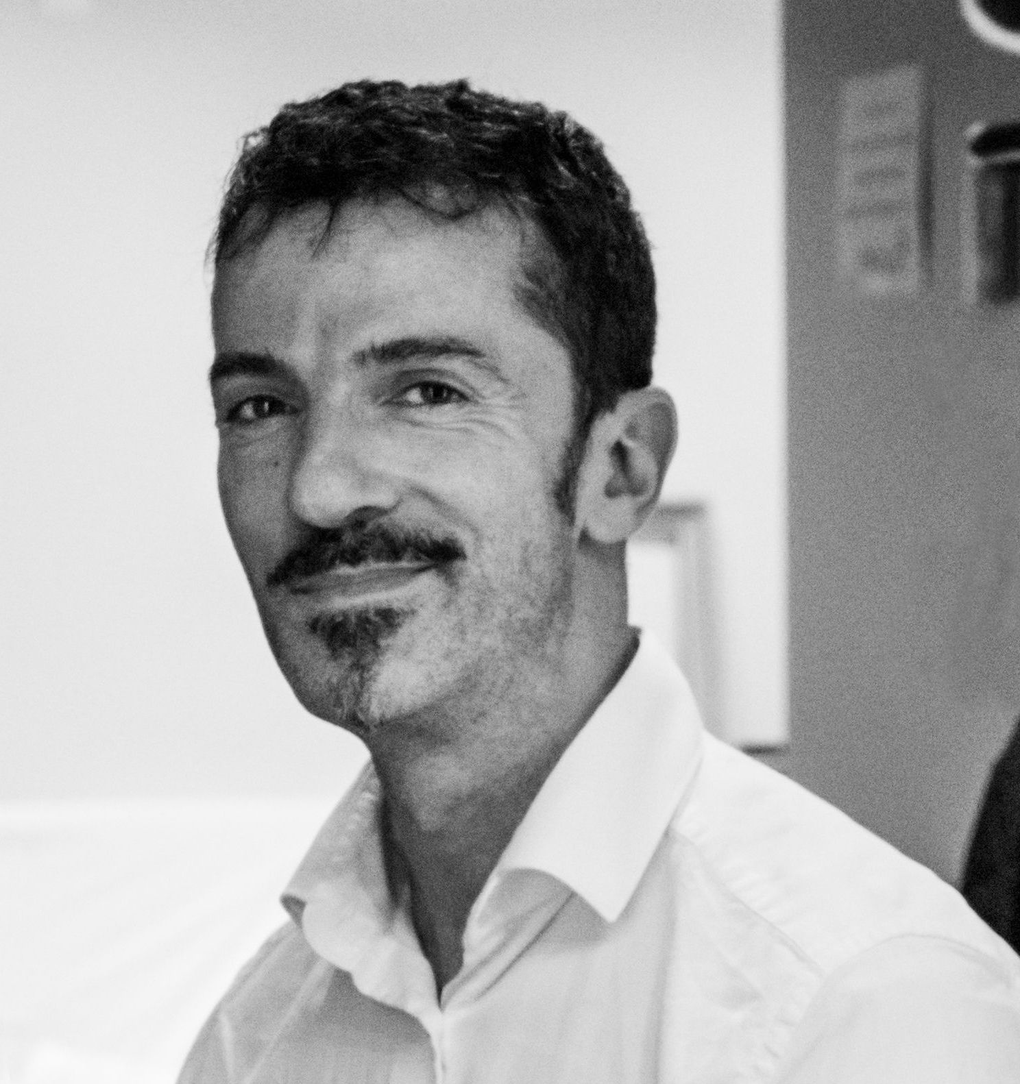 Man with dark hair and a goatee smiles, wearing a light-colored button-down shirt.