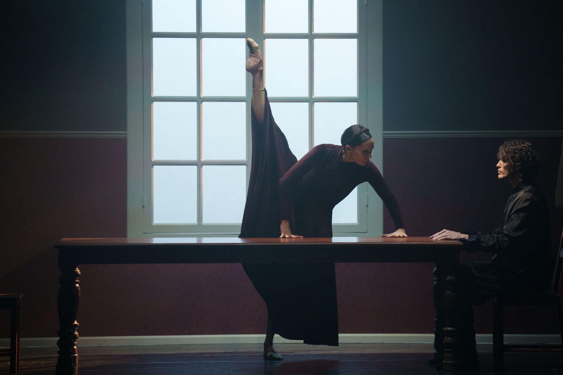 A person in a long dress does a split over a table, a second person seated opposite. Dark room with a window.