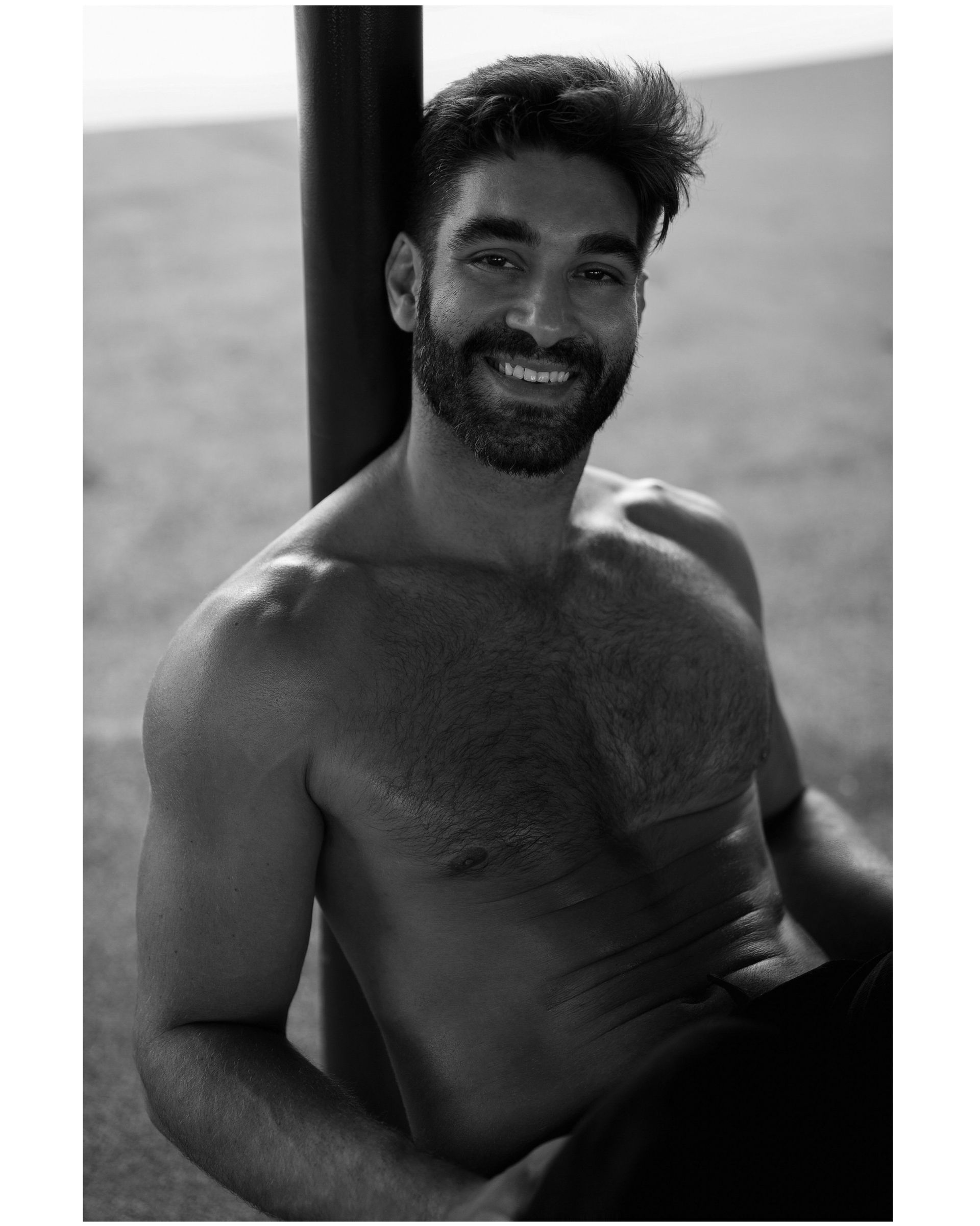 Shirtless man leaning against a pole, smiling. Outdoor setting.