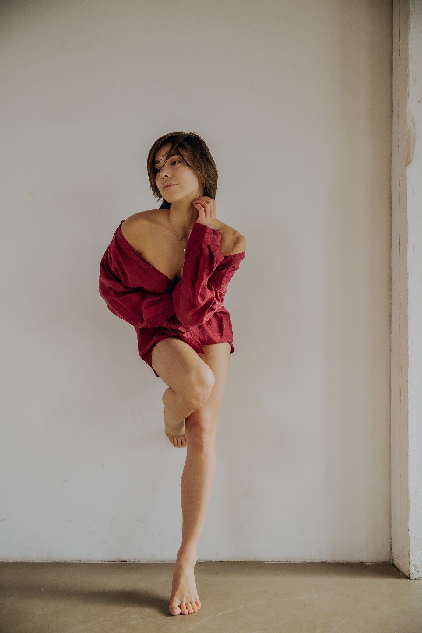 Woman in red shirt and shorts leaning against a white wall, leg crossed, barefoot.