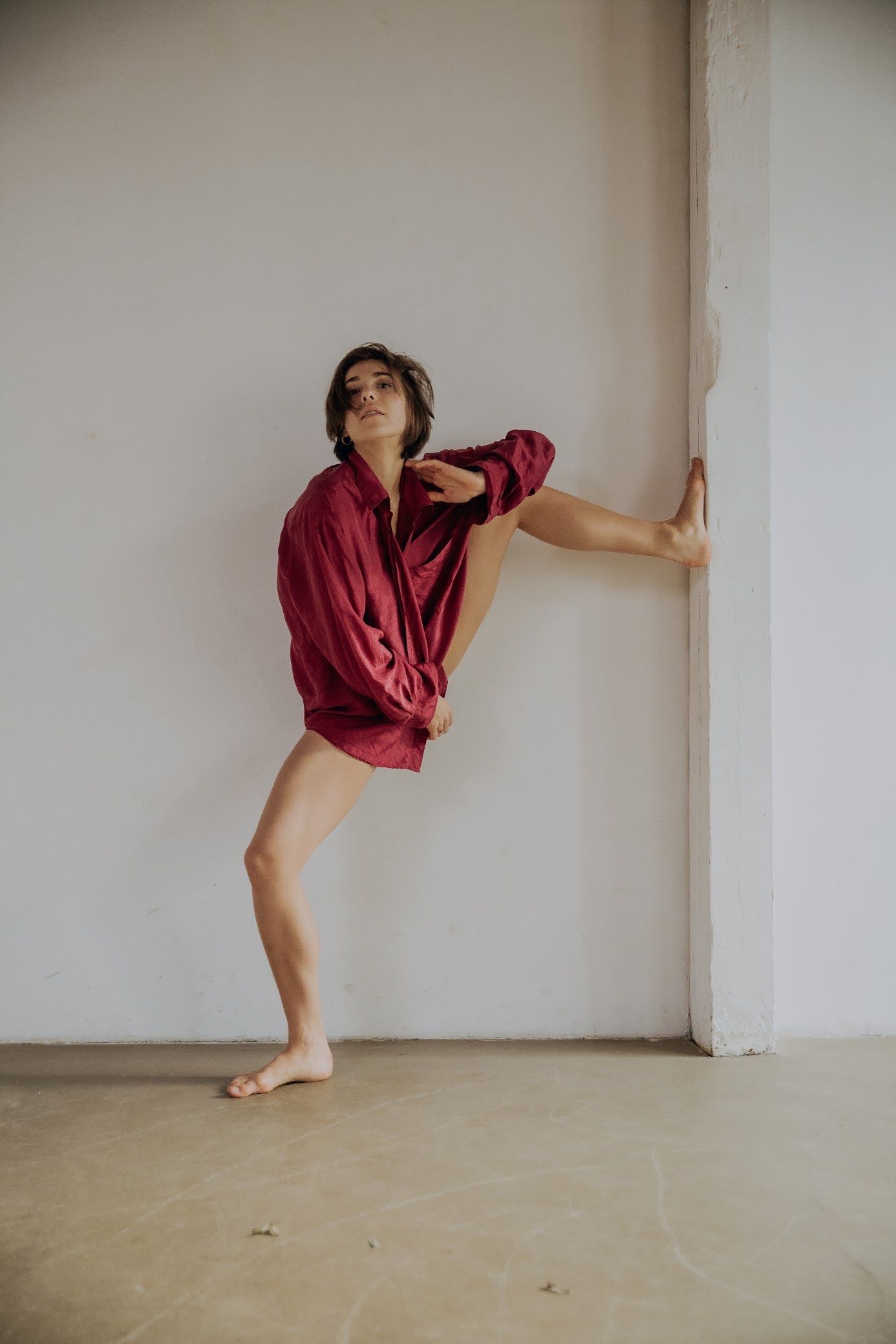 Person in a red oversized shirt, barefoot, doing a ballet stretch against a white wall and a pillar.