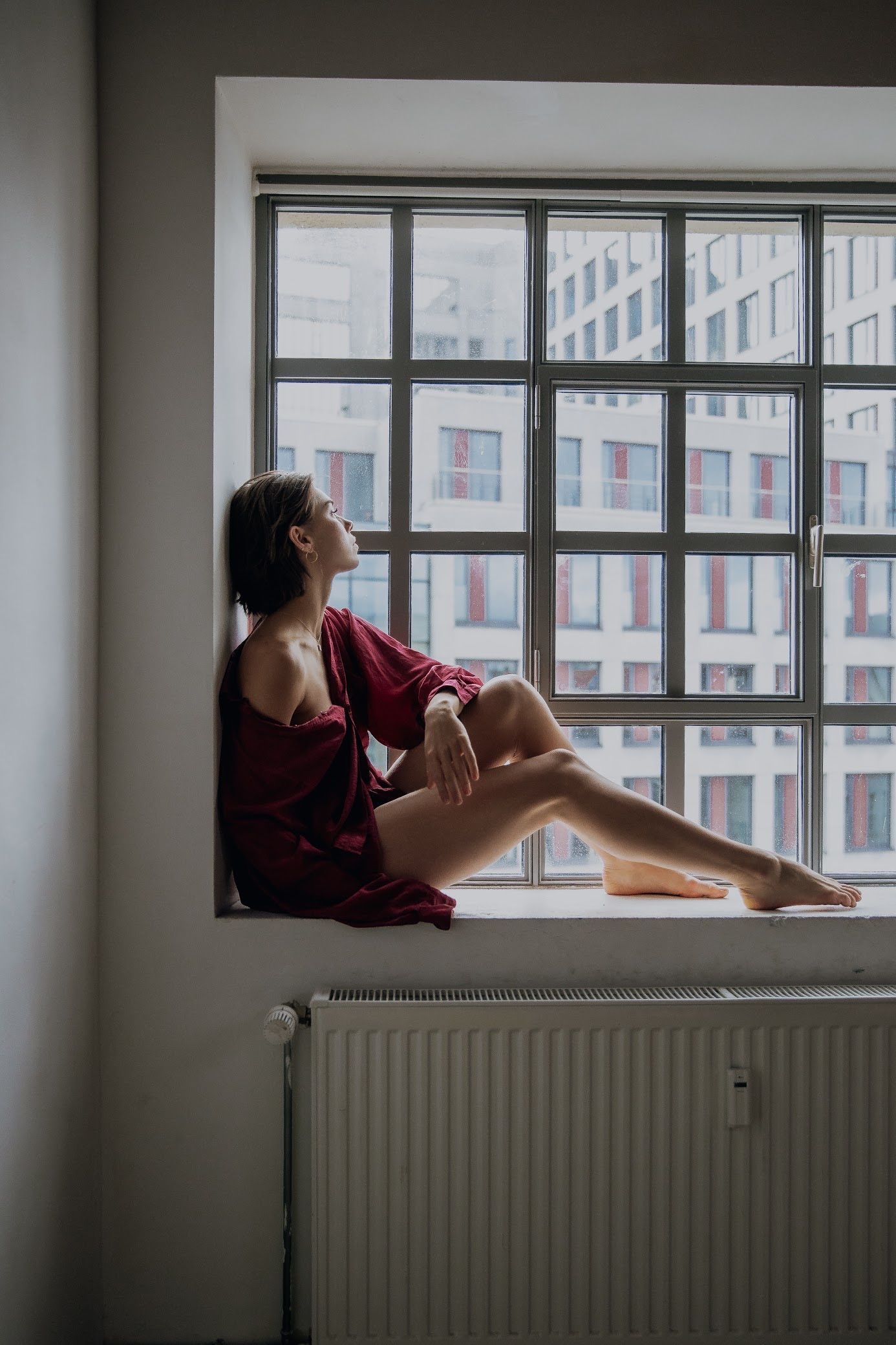 Woman in burgundy top sits in a window, looking up. City buildings visible through the panes.